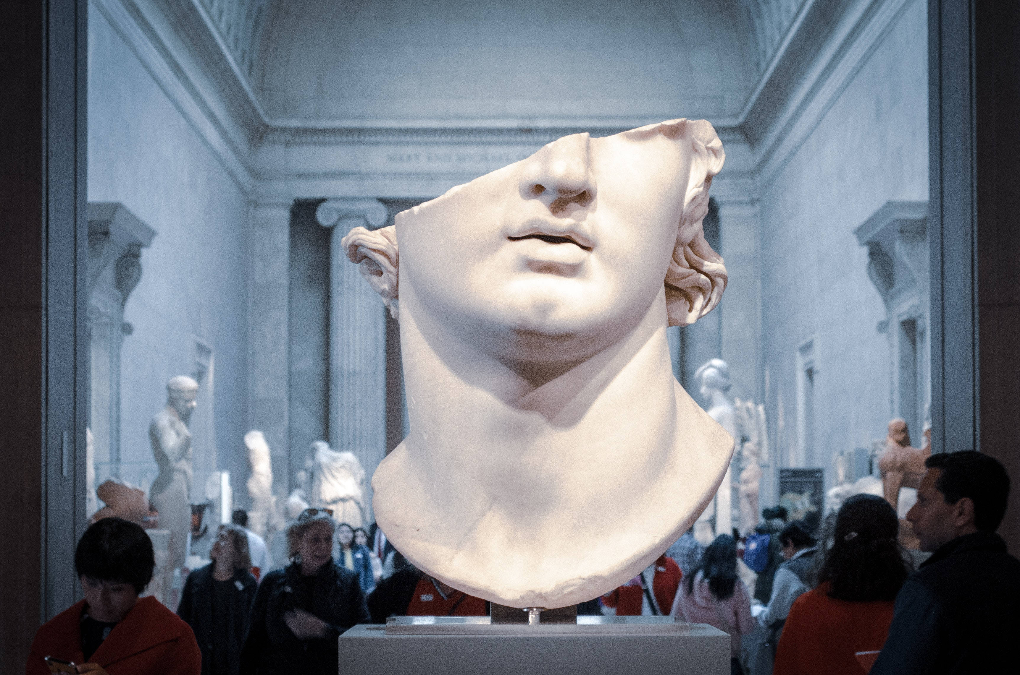 Sculpture 桌布 壁紙 — 一個缺少上半部臉部的大理石半身像，在一個充滿遊客和古典雕像的大型博物館畫廊中顯眼地矗立。