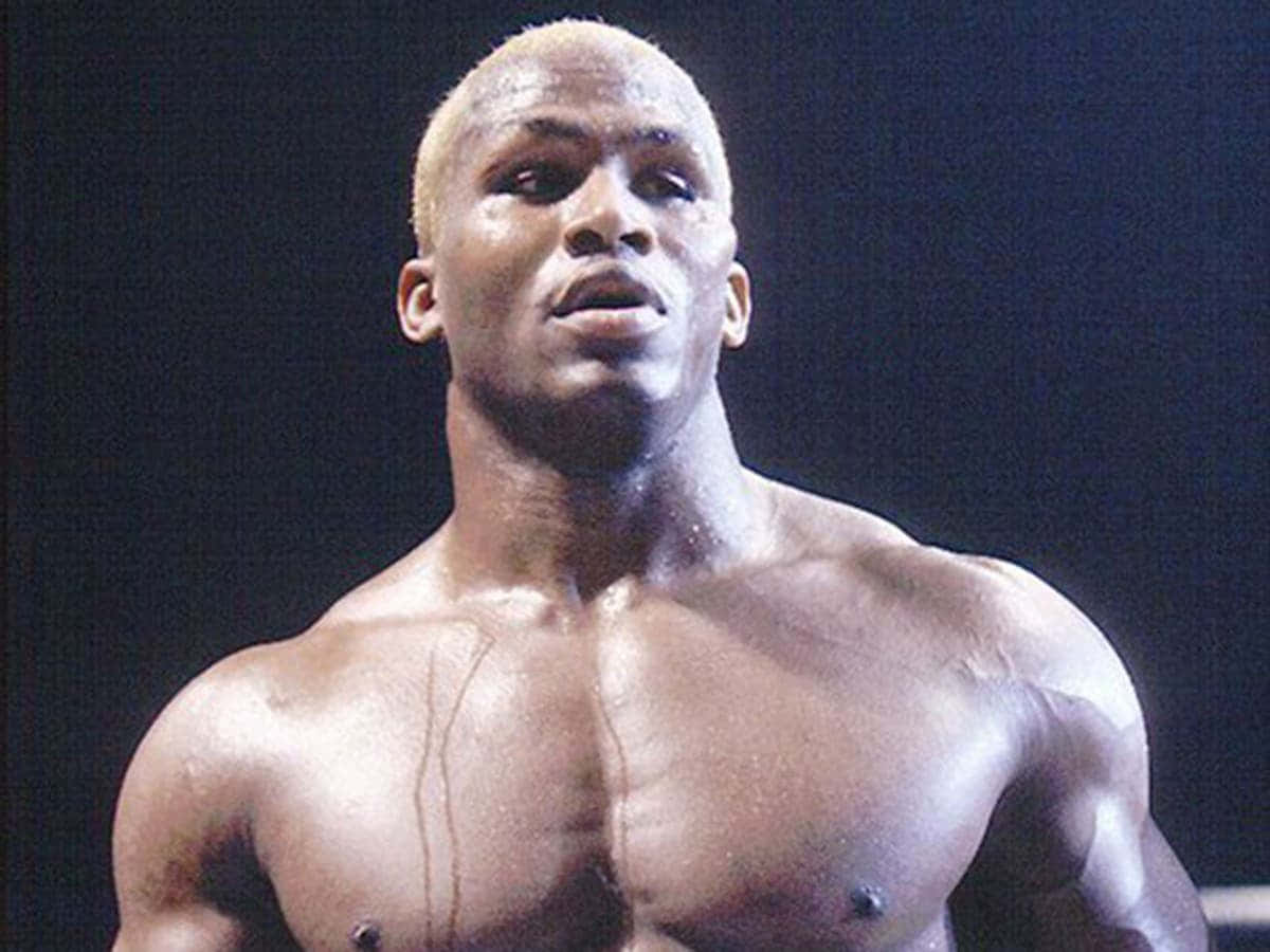 Kevin Randleman - Ultimate MMA Fighter 壁紙 — 一個肌肉發達、皮膚閃亮的摔角手在明亮的舞台燈光下，對著黑暗背景凝視。