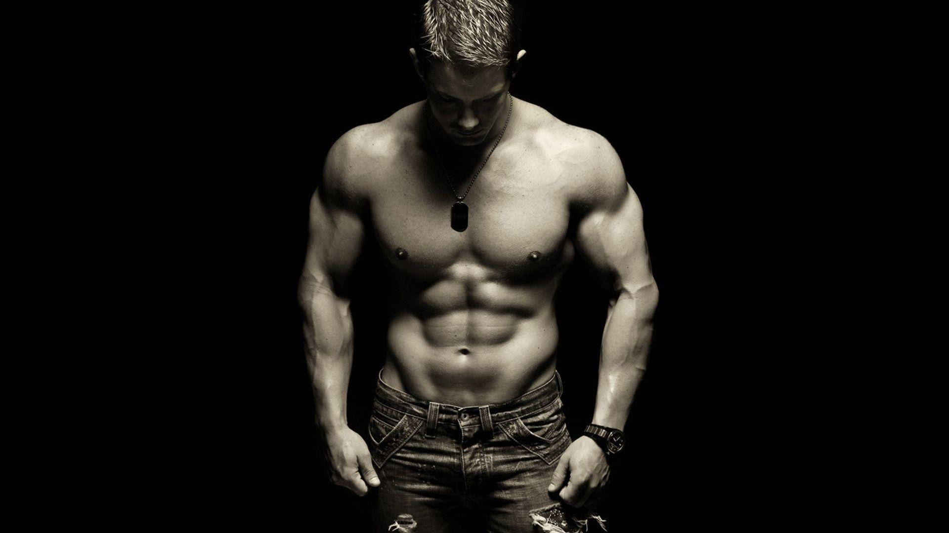 Shirtless Muscle Man Looking Down 壁紙 — 一個肌肉發達、腹肌分明的男人，赤裸上身，穿著牛仔褲和狗牌項鍊，站在黑色背景前。