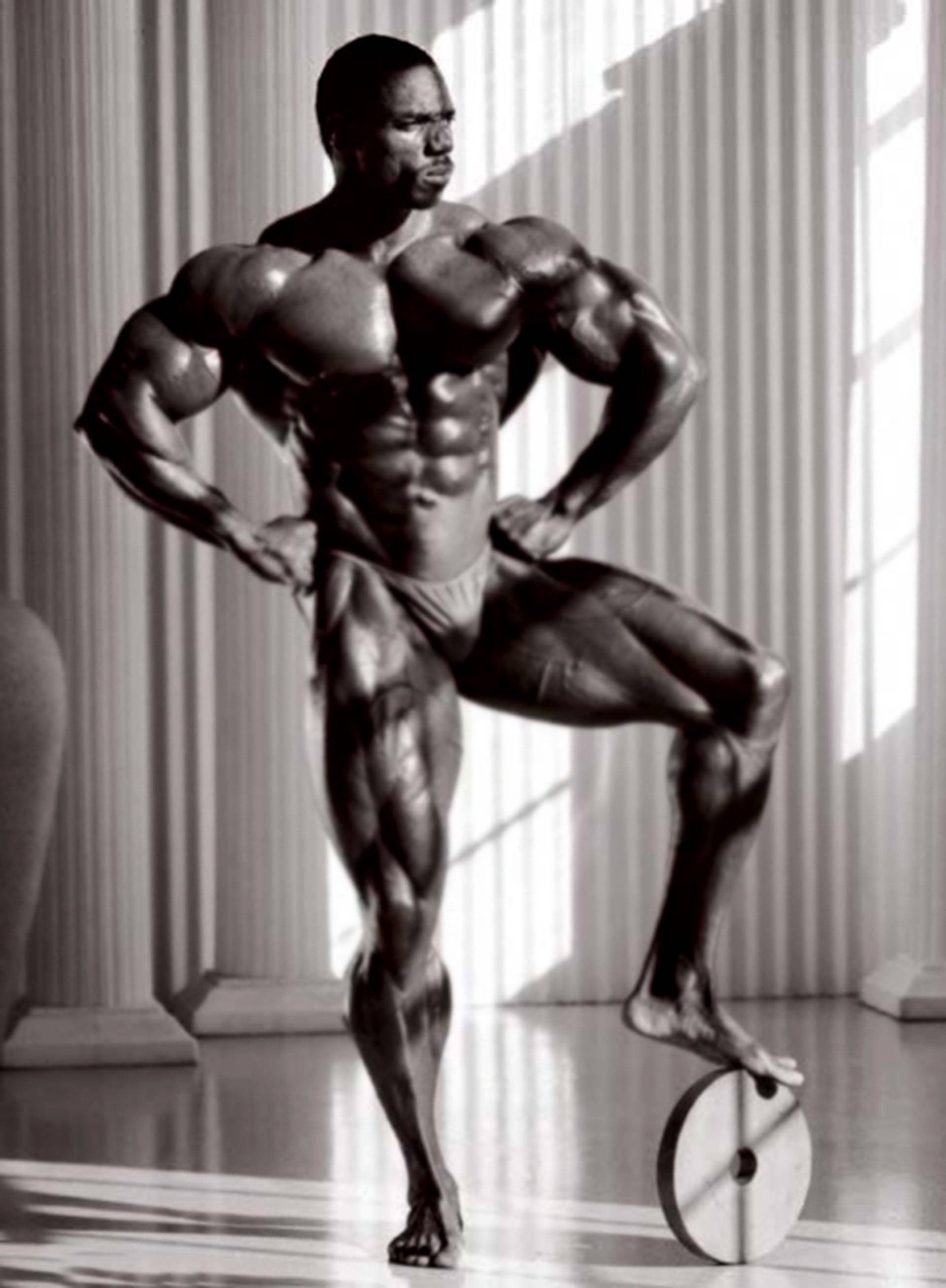 Herculean Flex Wheeler 壁紙 — 一個肌肉發達嘅男人喺單色調嘅工作室入面，帶住戲劇性燈光同古典柱，自信咁擺姿勢。