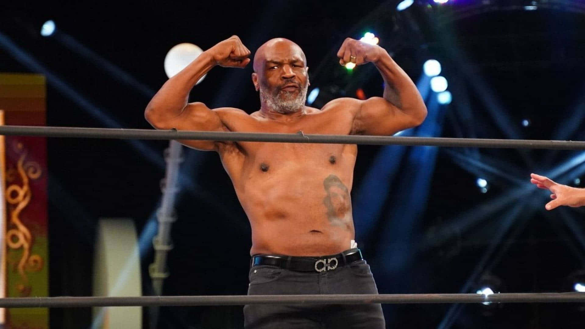 mike tyson 桌布 壁紙 — 一個肌肉發達的光頭摔角手在明亮的舞台燈光下於擂台上彎曲雙臂，腹部有紋身。