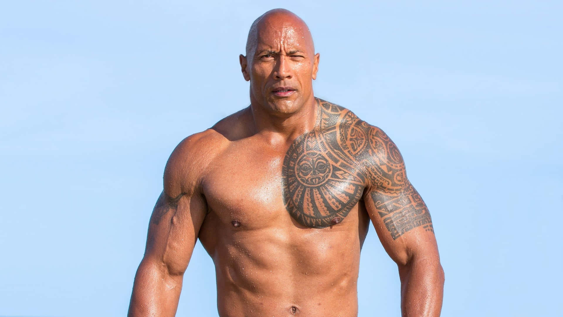 The Rock 桌布 壁紙 — 一個肌肉發達的光頭男子，胸口和手臂上有複雜的部落紋身，站在清澈的藍天下。