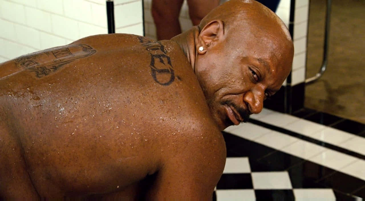 Ving Rhames 桌布 壁紙 — 一個肌肉發達的光頭男子，身上有紋身和戴著耳環，在瓷磚浴室裡向前傾，水滴在他皮膚上閃閃發光。