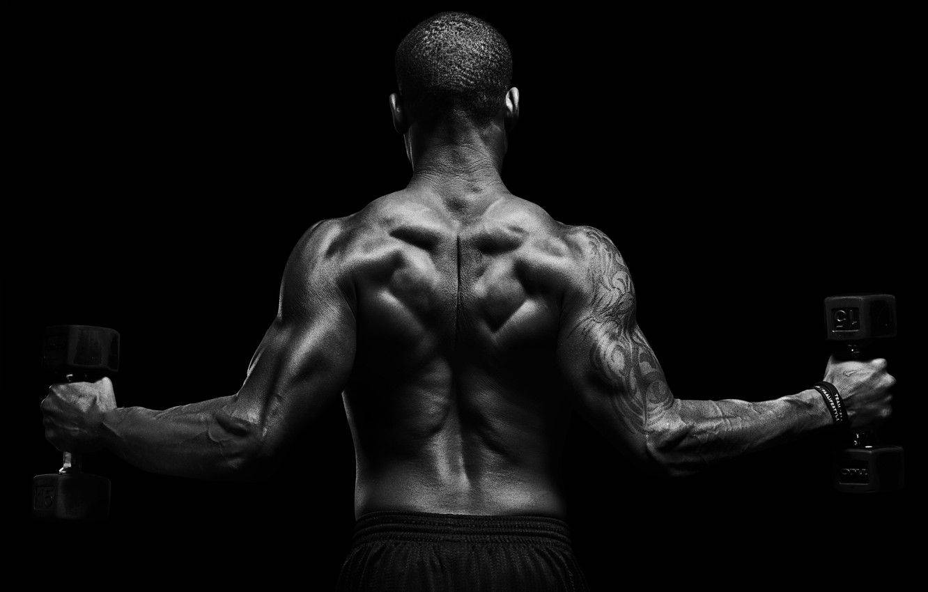 Muscle Man Flexing His Back 壁紙 — 一個肌肉發達的男人從背後觀看，在純黑色背景前舉起啞鈴，突顯他線條分明的背部和有紋身的手臂。