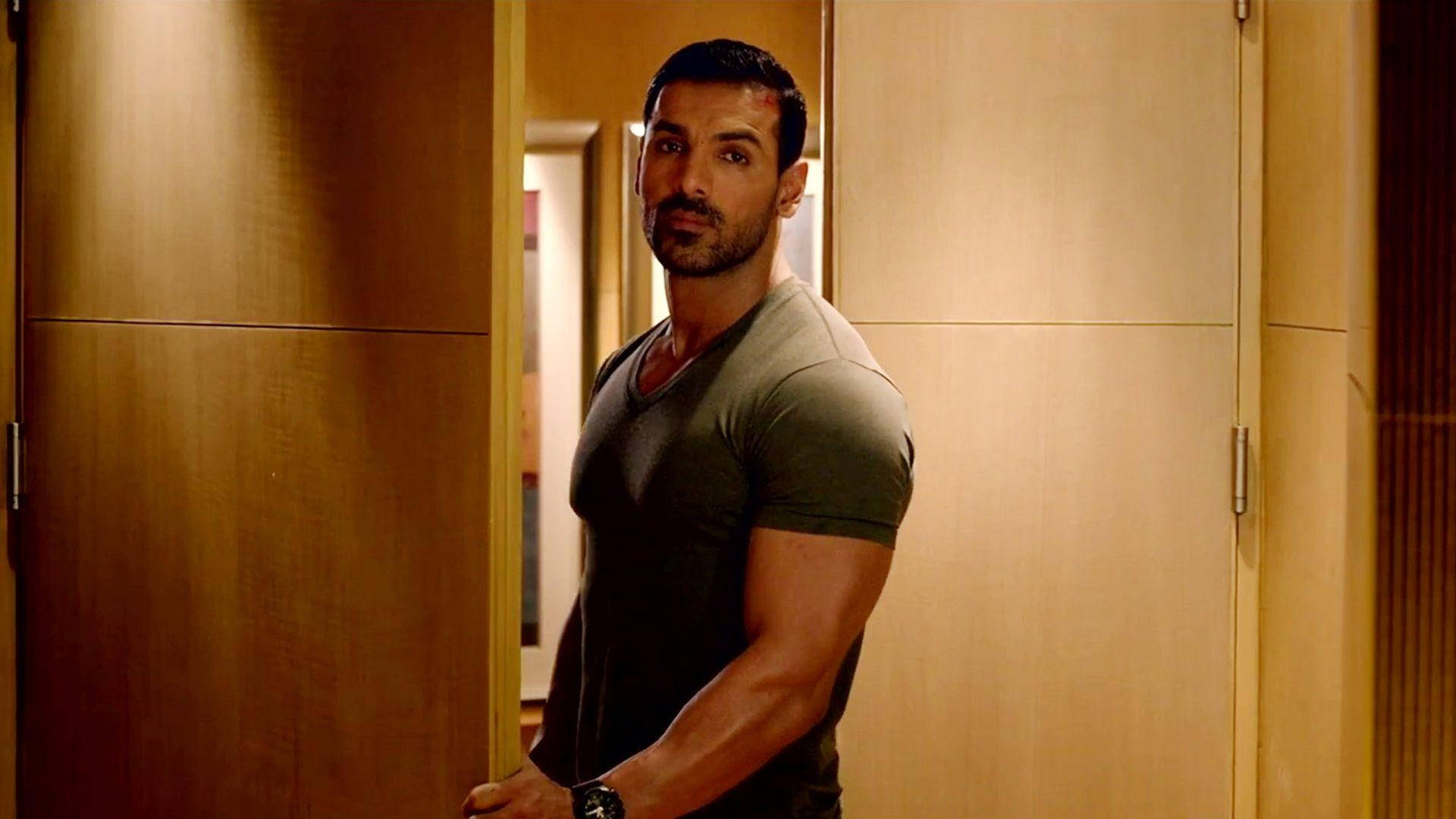 John Abraham HD 桌布 壁紙 — 一個肌肉發達的男人穿著灰色T恤，站在一個由木門框著的溫暖燈光走廊裡。