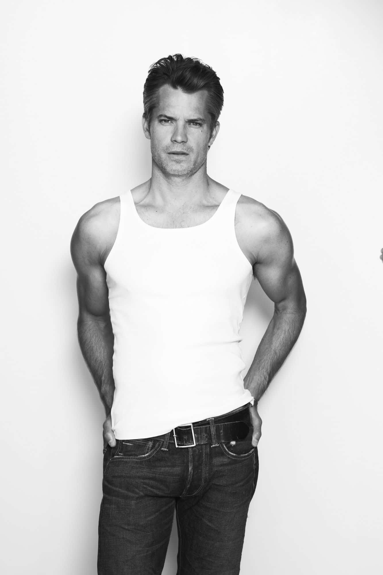 timothy olyphant 桌布 壁紙 — 一個肌肉發達的男人穿著白色背心和牛仔褲，在純白色背景前自信地站立，黑白肖像。