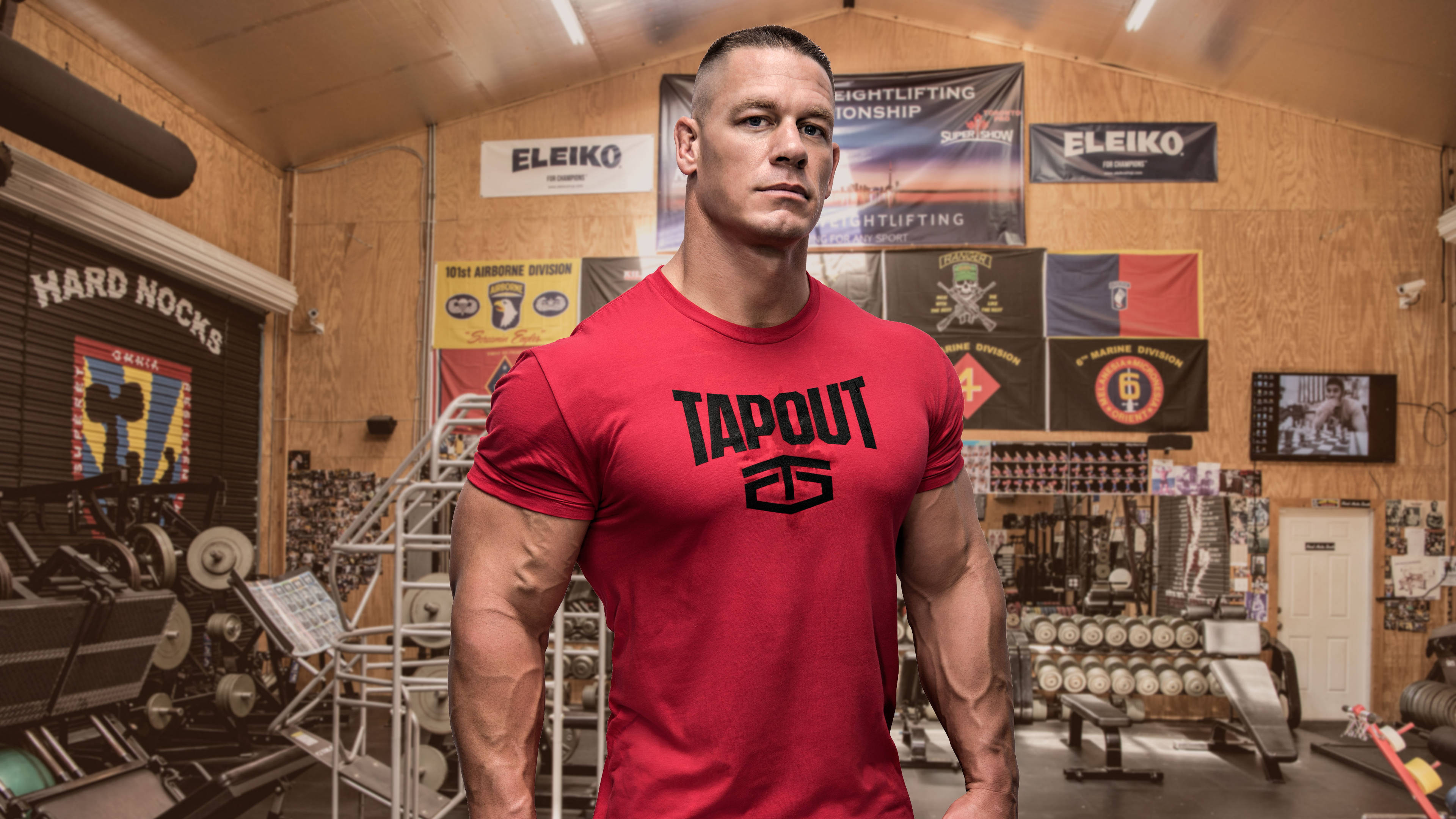 John Cena 桌布 壁紙 — 一個肌肉發達的男人穿著紅色Tapout襯衫，自信地站在一個雜亂的健身房裡，裡面充滿了舉重設備和勵志橫幅。