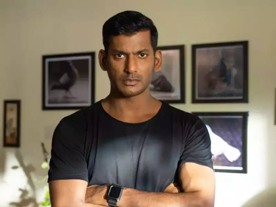 Indian 桌布 壁紙 — 一個肌肉發達的男人穿著黑色T恤，在一個光線柔和、身後牆上掛著裱框照片的房間裡交叉雙臂站立。