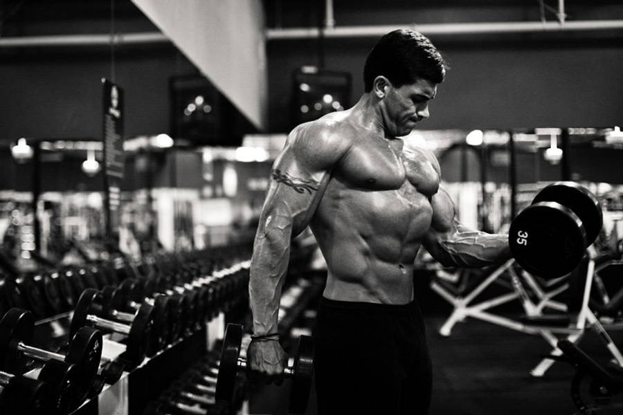 Exercise 桌布 壁紙 — 一個肌肉發達的男子在燈光昏暗的健身房中用力舉起啞鈴，背景有啞鈴架。