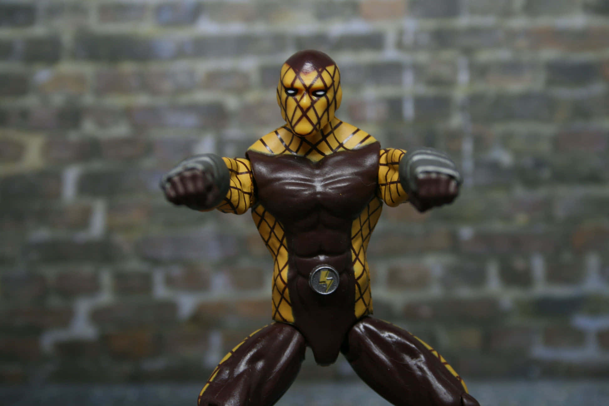 The Shocker 桌布 壁紙 — 一個肌肉發達的黃色和棕色裝甲動作人偶，以戰鬥姿勢站立在模糊的磚牆背景前。