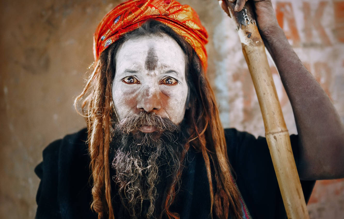 Aghori 桌布 壁紙 — 一個臉上塗有白色顏料、戴著橙色頭巾的男人，手持木杖靠著紋理牆壁。
