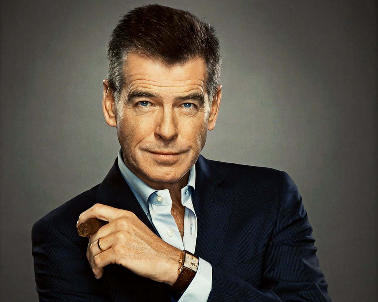pierce brosnan 桌布 壁紙 — 一個自信嘅男人着住深色西裝，喺柔和灰色背景前揸住雪茄，有強烈嘅藍眼睛。