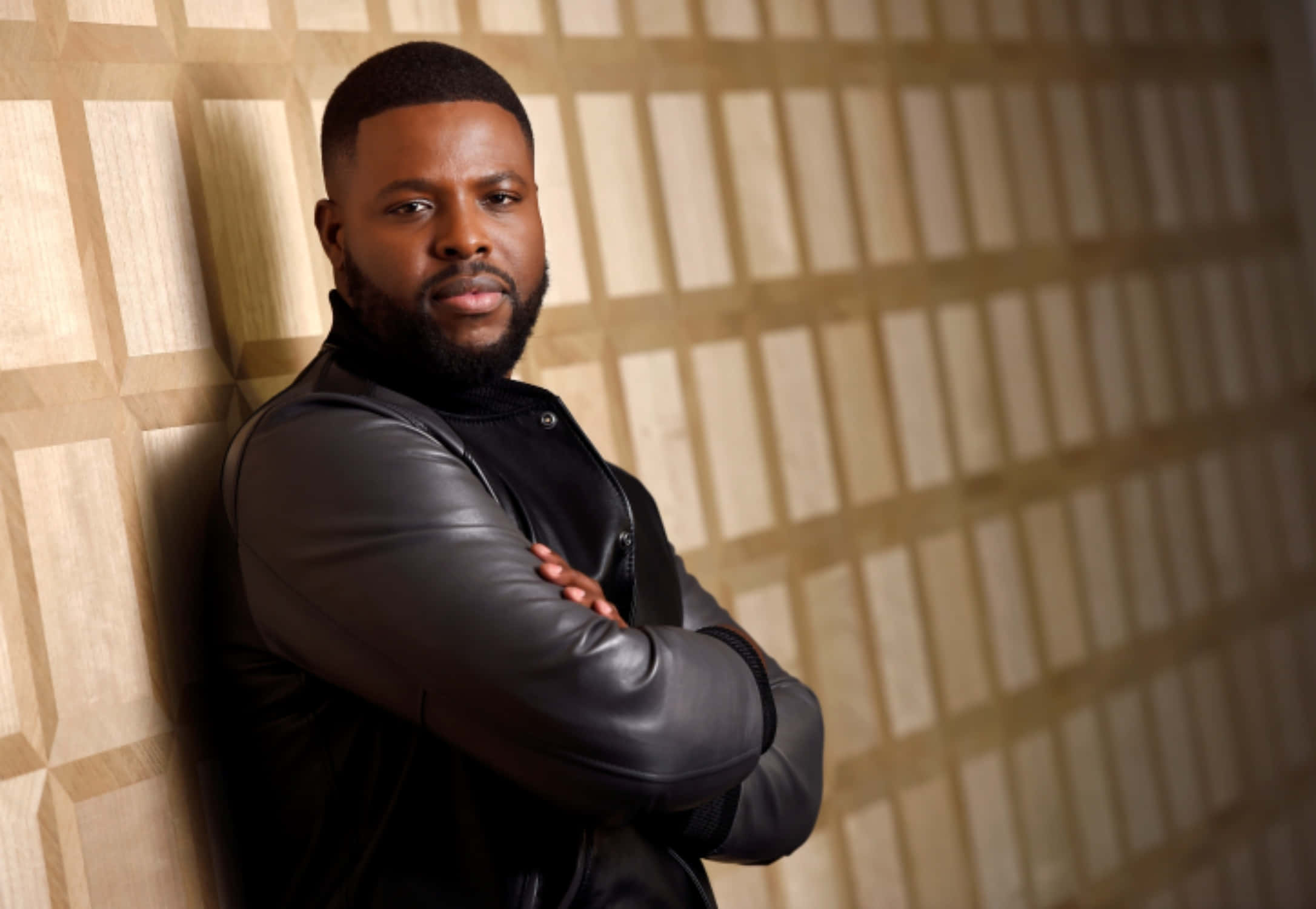 Winston Duke 桌布 壁紙 — 一個自信嘅鬍鬚男人靠喺幾何圖案牆上，著住黑色皮褸。