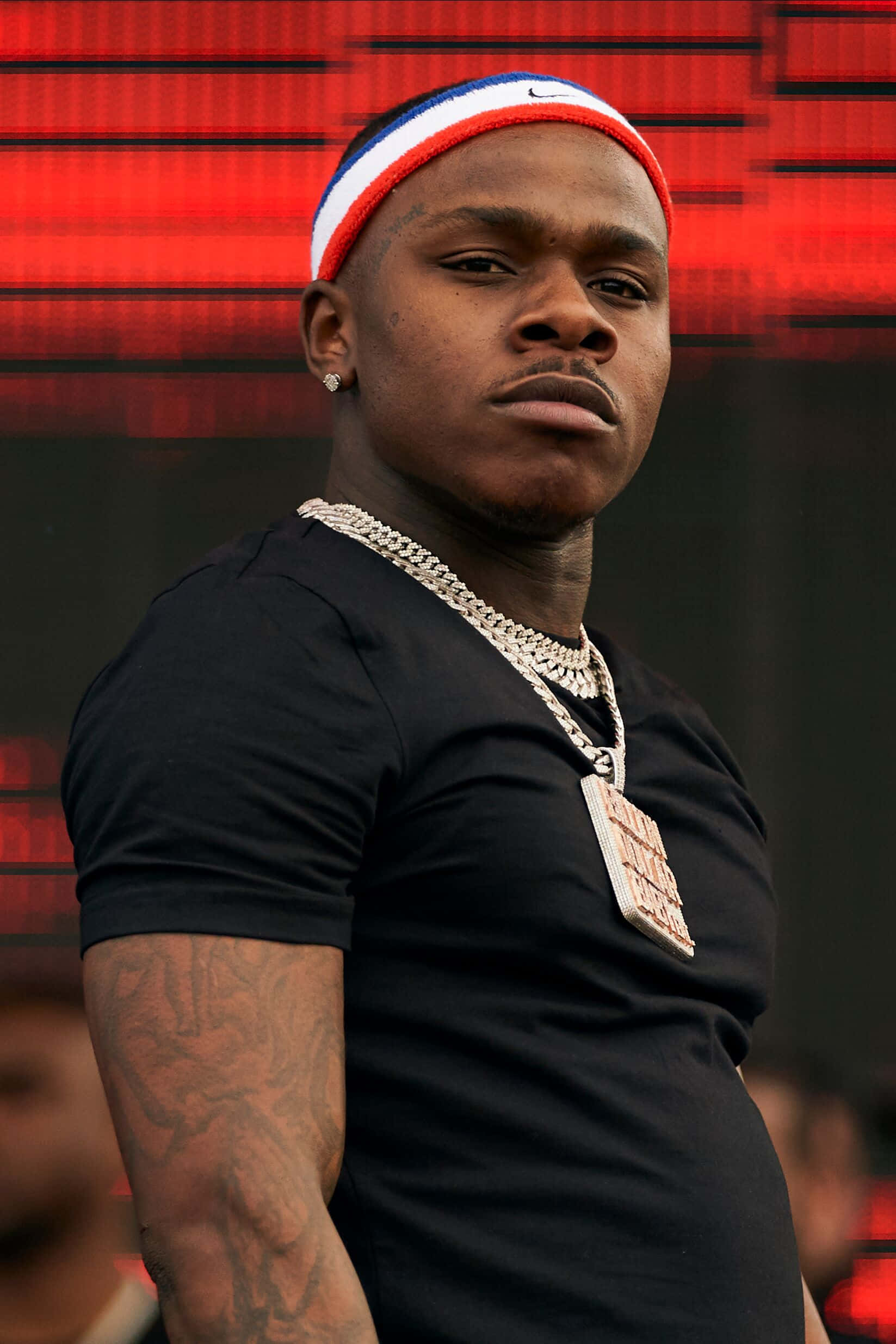 Dababy 桌布 壁紙 — 一個自信的男人，手臂上有紋身，戴著紅白藍頭帶和粗鏈條項鍊，背景是發光的紅色數位背景。