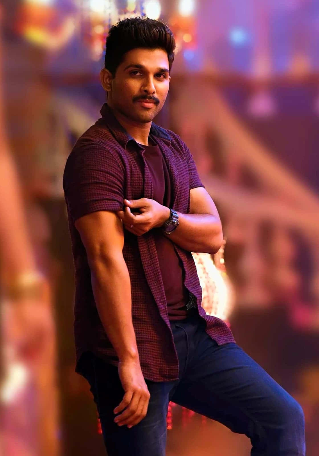 Allu Arjun 桌布 壁紙 — 一個自信的男人穿著栗色格仔襯衫，一手放在手臂上，在充滿活力的模糊舞台背景前擺姿勢。