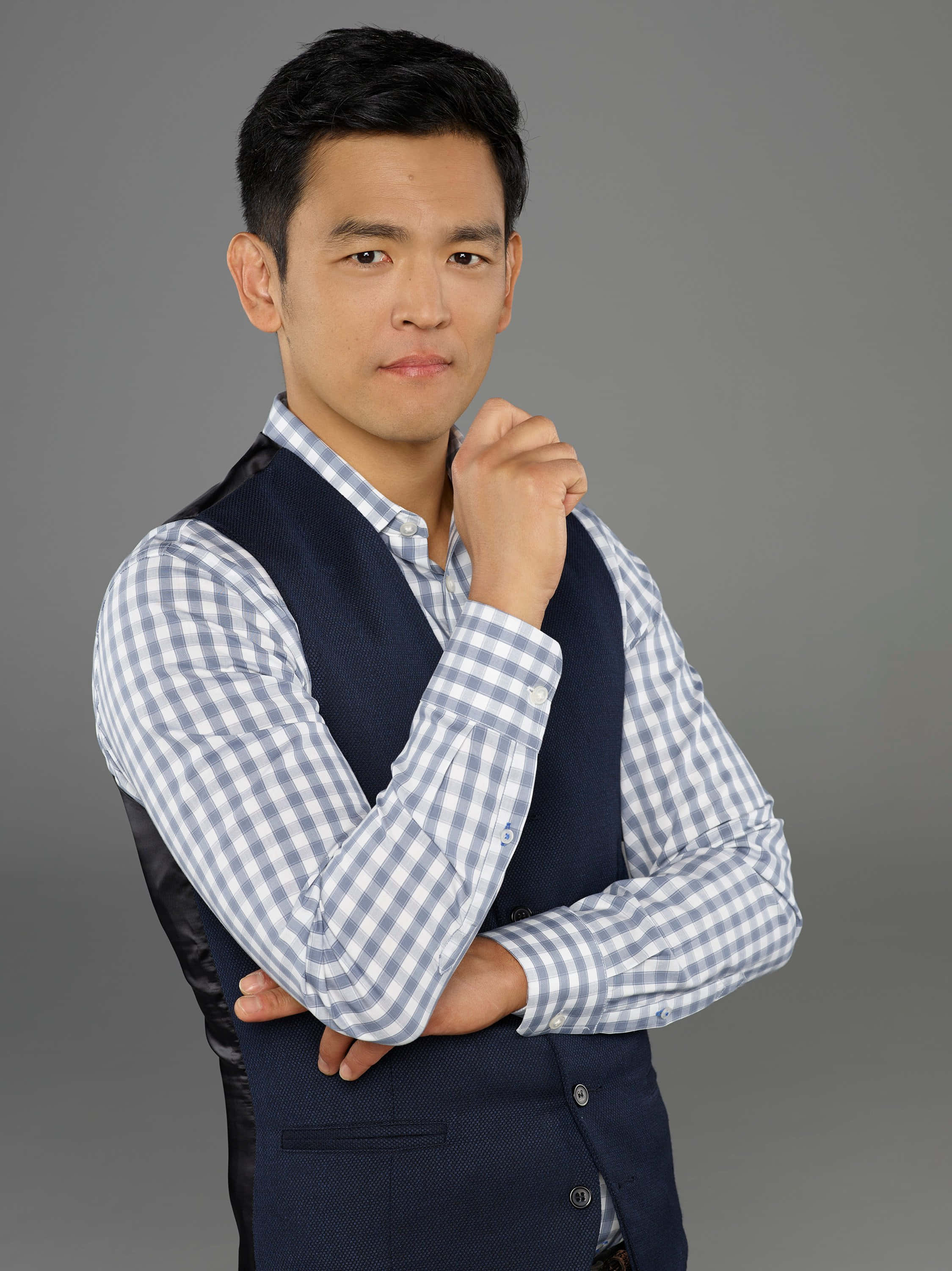 John Cho 桌布 壁紙 — 一個自信的男人穿著藍色背心和格仔襯衫，在純灰色背景前將拳頭靠近下巴擺姿勢。