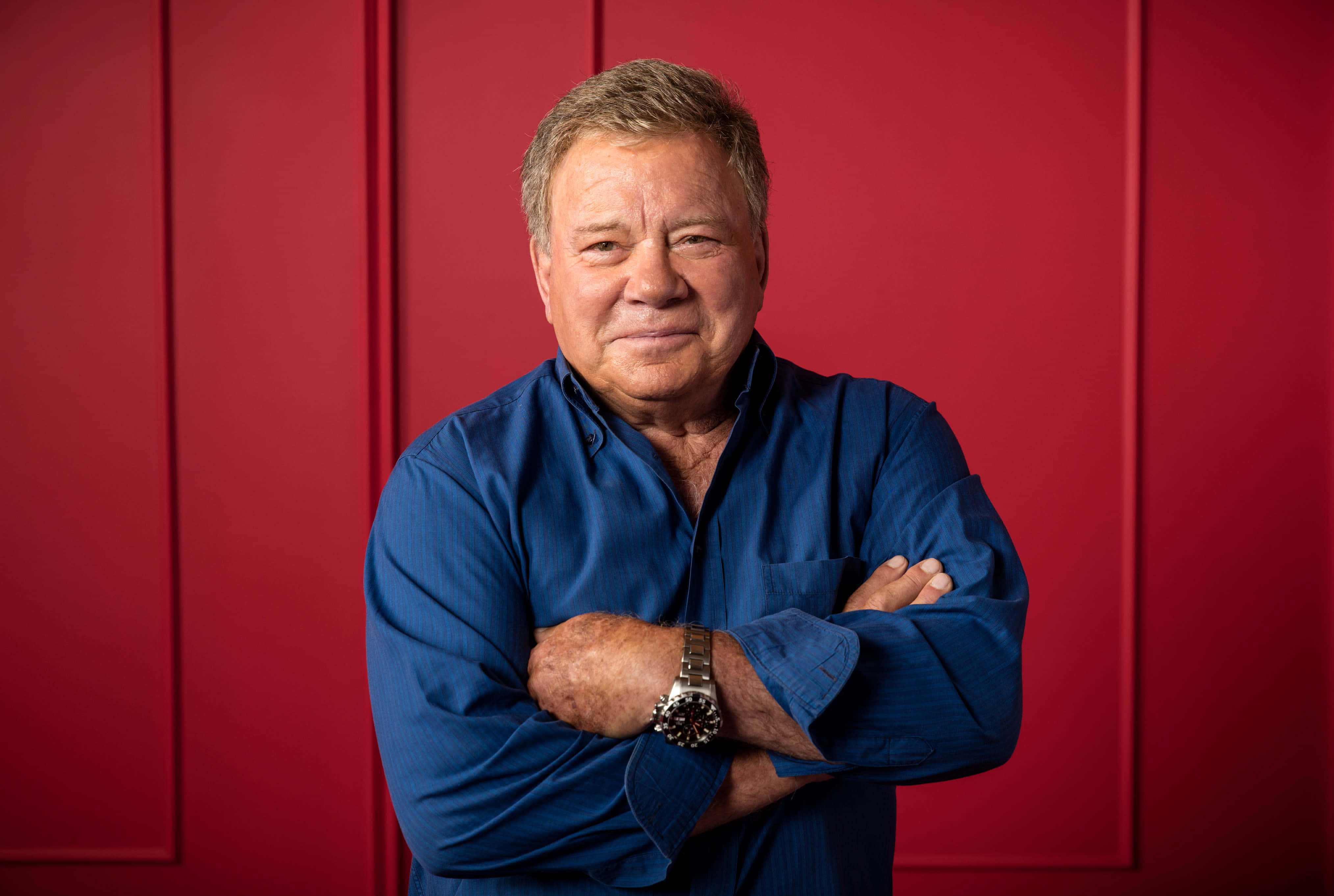 william shatner 桌布 壁紙 — 一個自信的男人穿著藍色襯衫，雙臂交叉站在大膽的紅色鑲板牆前，戴著銀色手錶。
