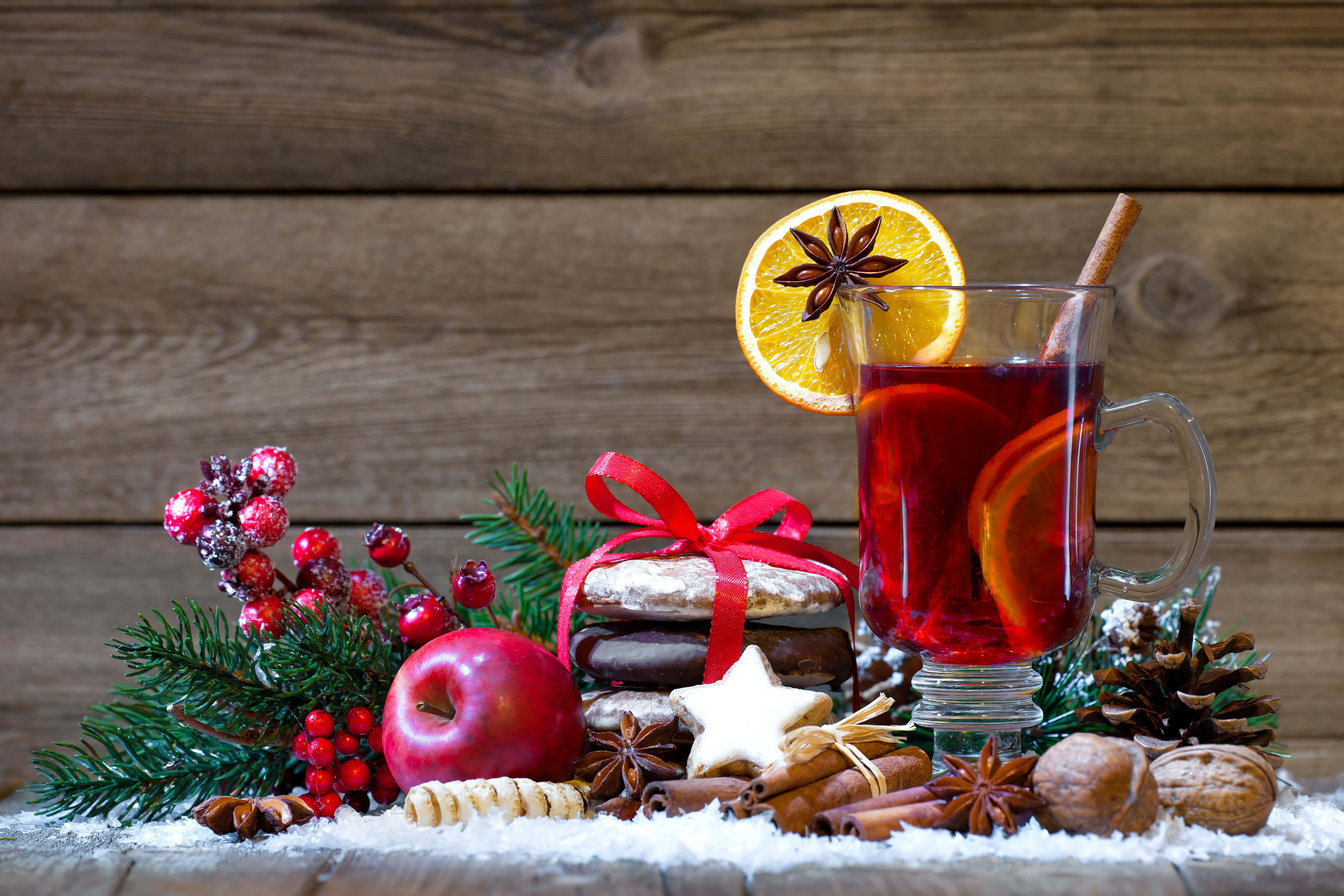 Mulled Wine 桌布 壁紙 — 一個舒適的冬季假日場景，一杯加了香料的热红酒，以橙子和八角裝飾，旁邊有節日餅乾、松果和紅莓，放在雪覆蓋的木頭上。