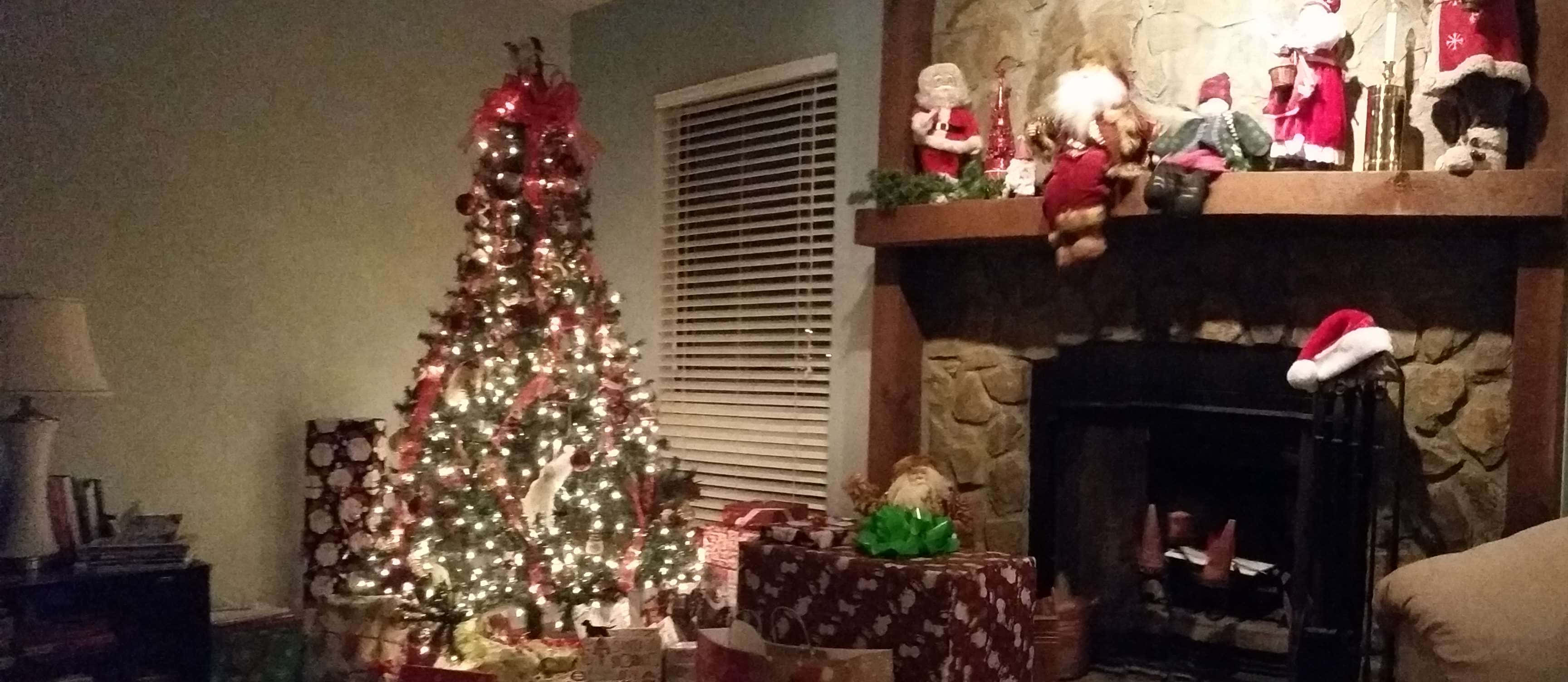 Rustic Christmas 桌布 壁紙 — 一個舒適的客廳，有一棵點亮的聖誕樹，裝飾著紅色絲帶和禮物，旁邊是一個石頭壁爐，裝飾著聖誕老人公仔和聖誕帽。