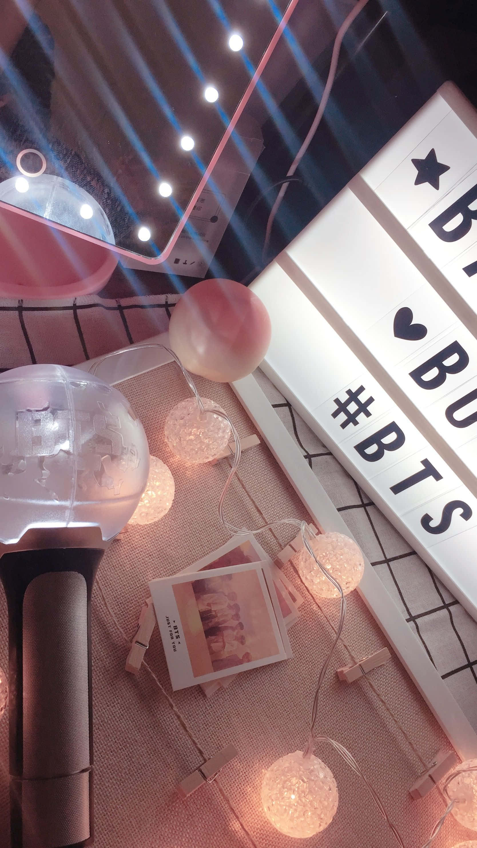 Bts Army Bomb 桌布 壁紙 — 一個舒適的室內場景，粉紅色串燈在BTS燈箱和寶麗來照片旁邊溫暖地發光，放在格子表面上。
