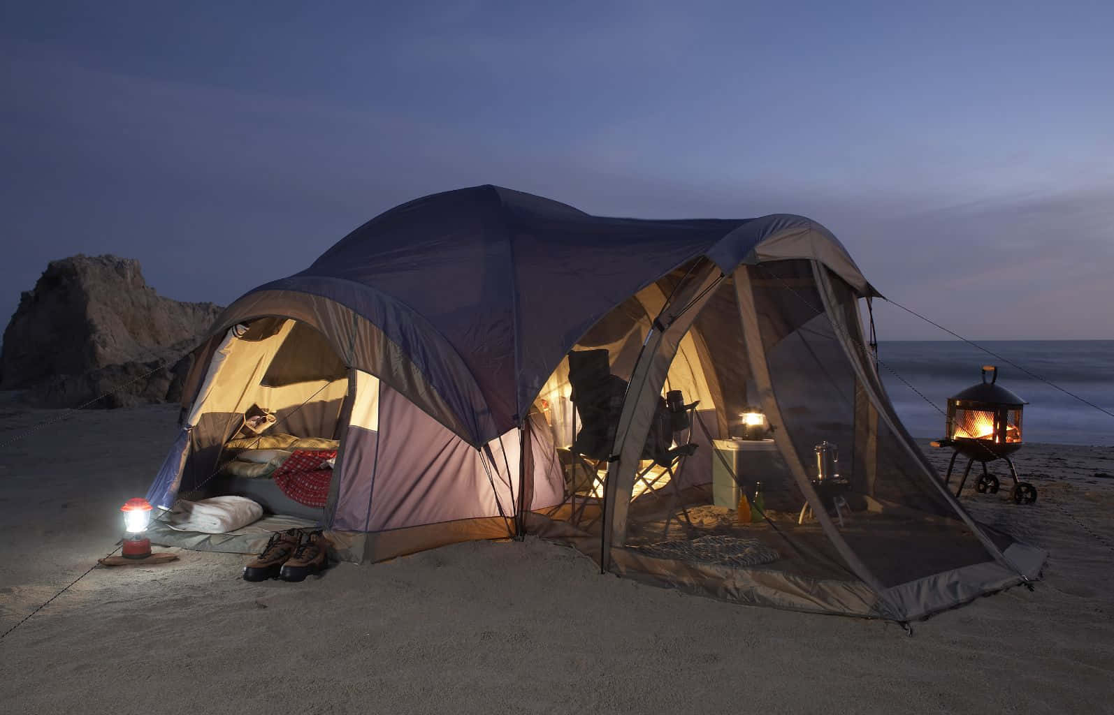 Beach Camping 桌布 壁紙 — 一個舒適的帳篷在黃昏的沙灘上溫暖地發光，營火和燈籠照亮了內部及周圍的岩石。
