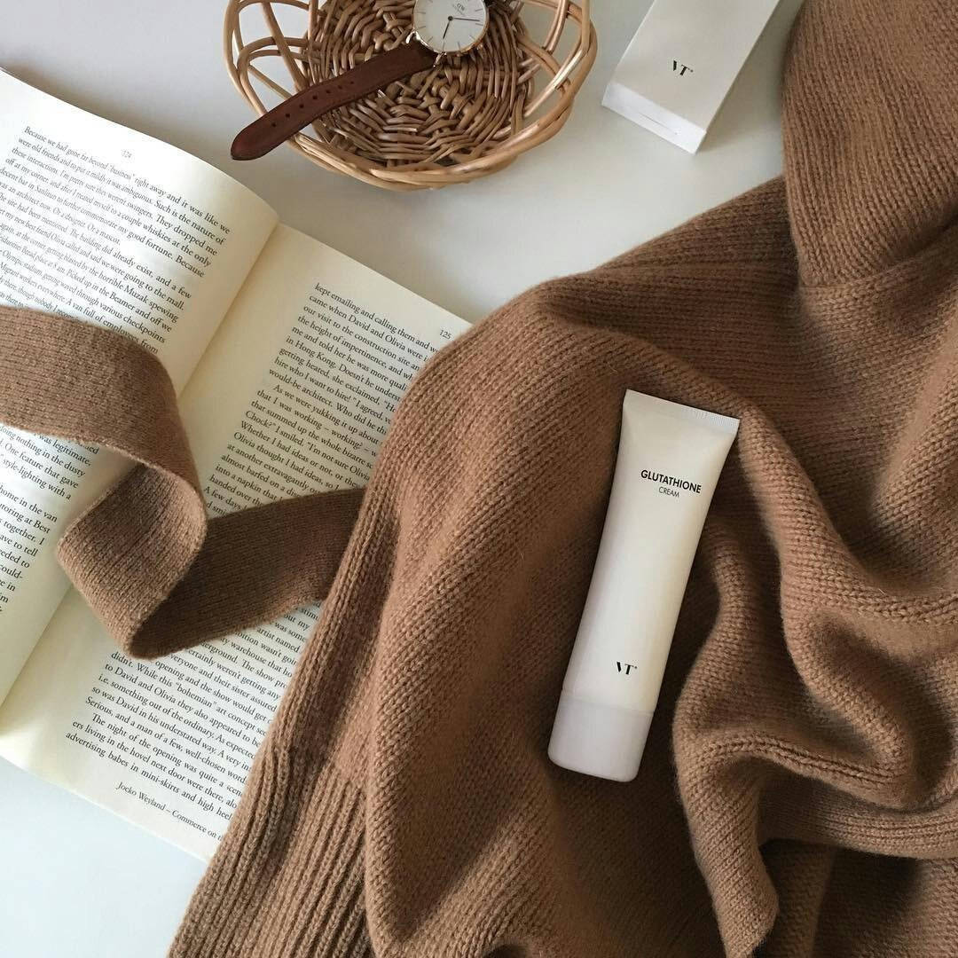 Beige Minimalist 桌布 壁紙 — 一個舒適的平鋪構圖，包括棕色針織毛衣、打開的書本、編織籃內的手錶，以及白色穀胱甘肽面霜，置於白色表面上。