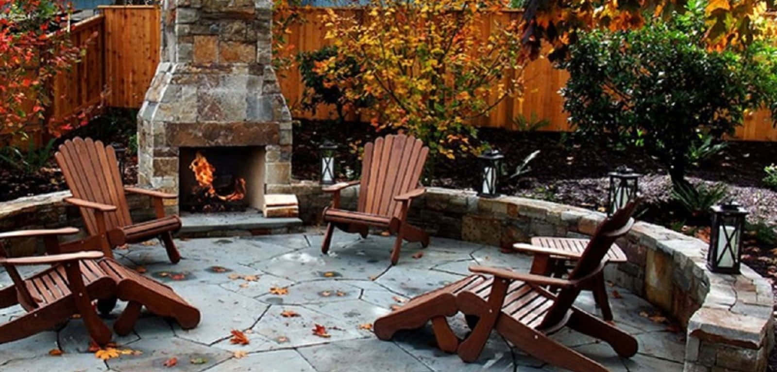 Autumn Fireplace 桌布 壁紙 — 一個舒適的戶外石壁爐，周圍環繞著木製 Adirondack 椅子和飄落的秋葉。
