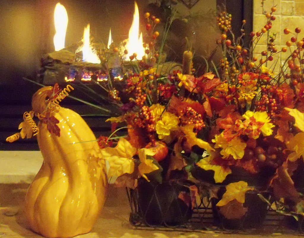Autumn Fireplace 桌布 壁紙 — 一個舒適的秋日場景，壁爐在鮮豔的花藝擺設和一個黃色陶瓷葫蘆後方閃爍著光芒。