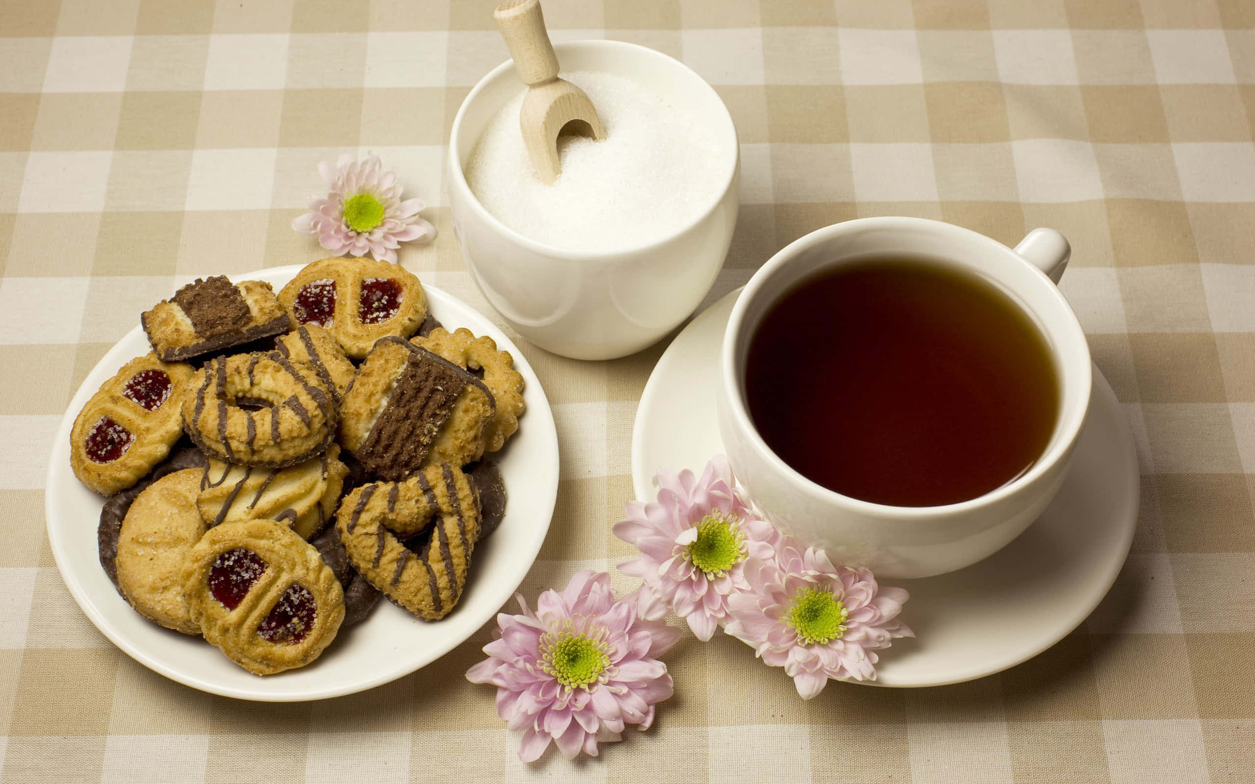 Floral Tea 桌布 壁紙 — 一個舒適的茶飲擺設，包括各式餅乾、一杯濃茶和糖碗，擺放在帶有粉紅色花朵的格仔枱布上。