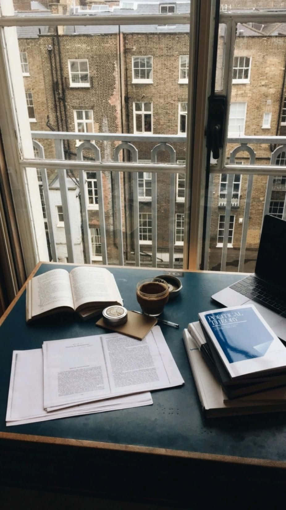 溫習 桌布 壁紙 — 一個舒適的讀書角落，暗色書桌上放著打開的書籍、紙張和一杯咖啡，透過玻璃門俯瞰磚砌建築。