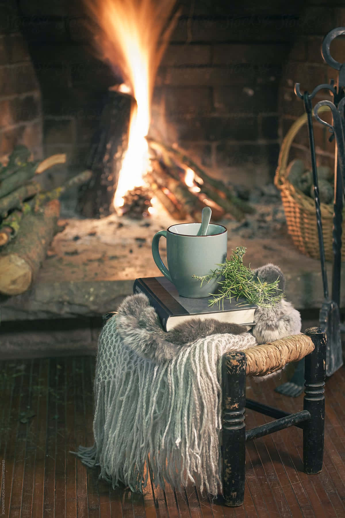 Cozy Fire 桌布 壁紙 — 一個舒適的馬克杯和一本書放在一個鋪著流蘇毯子的鄉村風格凳子上，前面是噼啪作響的壁爐。