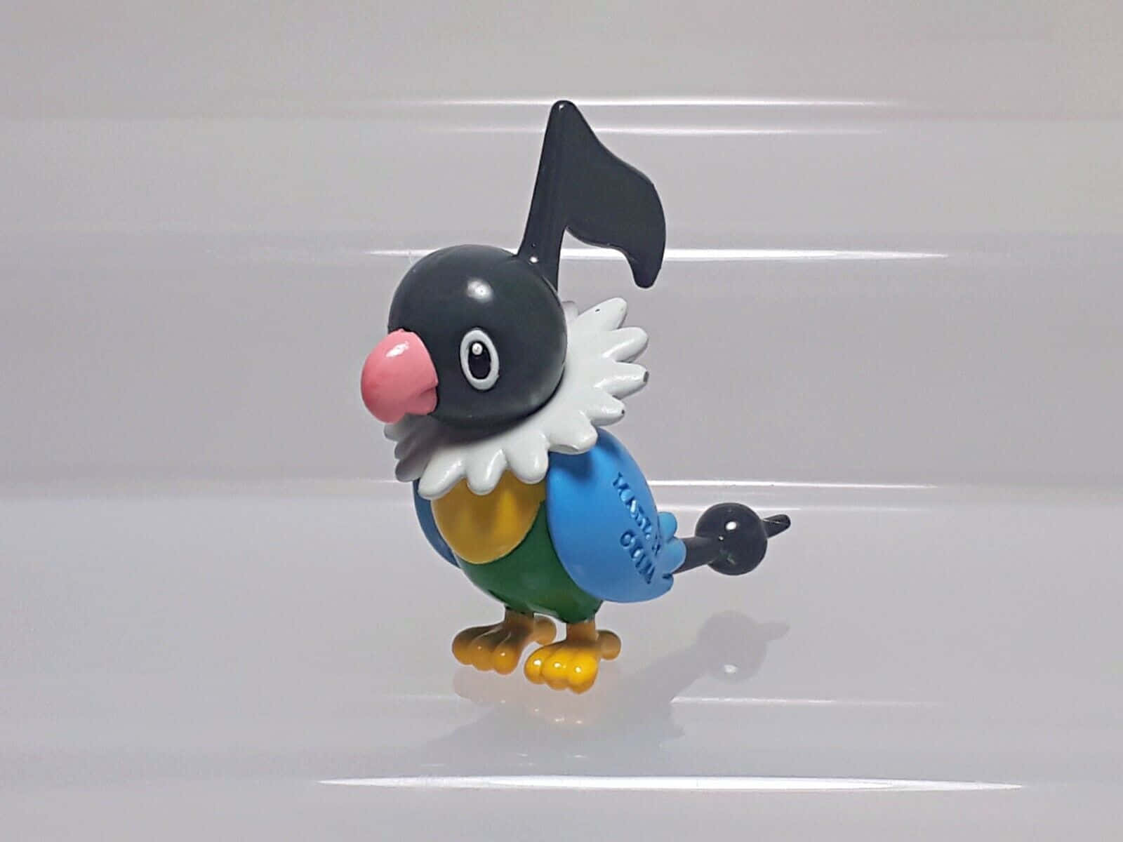 Pokemon Figures 桌布 壁紙 — 一個色彩繽紛的寶可夢Oricorio塑膠公仔，站在帶有黑色音符冠飾的光滑白色表面上。