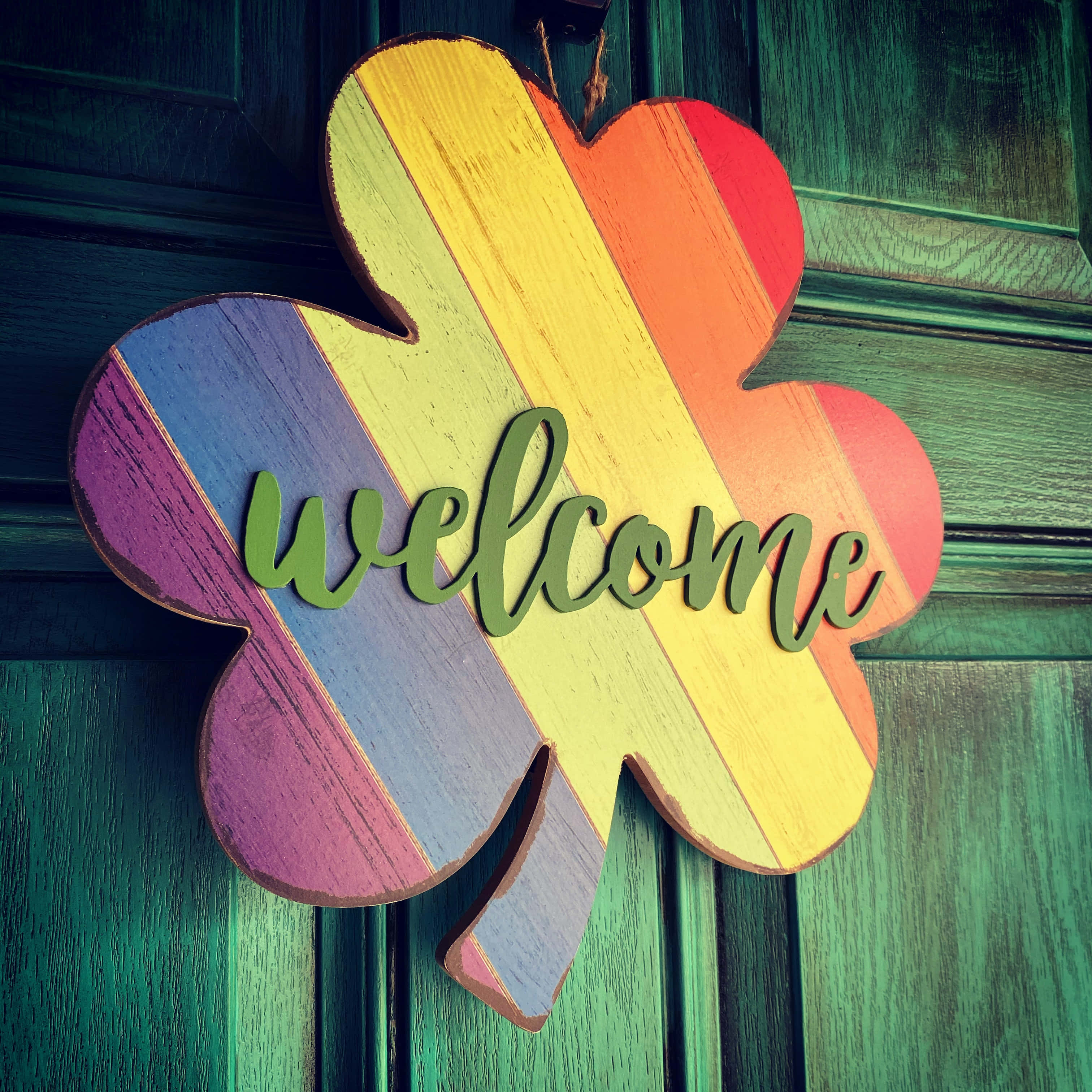 Welcome 桌布 壁紙 — 一個色彩繽紛的彩虹色三葉草門掛，上面用綠色字體寫著「歡迎」，掛在一扇風化的青綠色木門上。