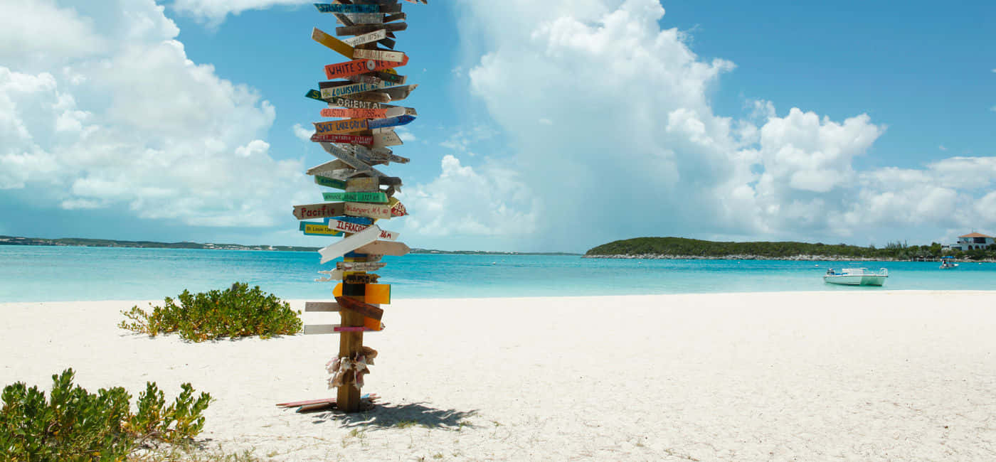 Bahamas Island 桌布 壁紙 — 一個色彩繽紛的木製路標矗立在純淨的白色沙灘上，沙灘上有碧綠的海水和遠處的島嶼，天空湛藍，雲朵蓬鬆。