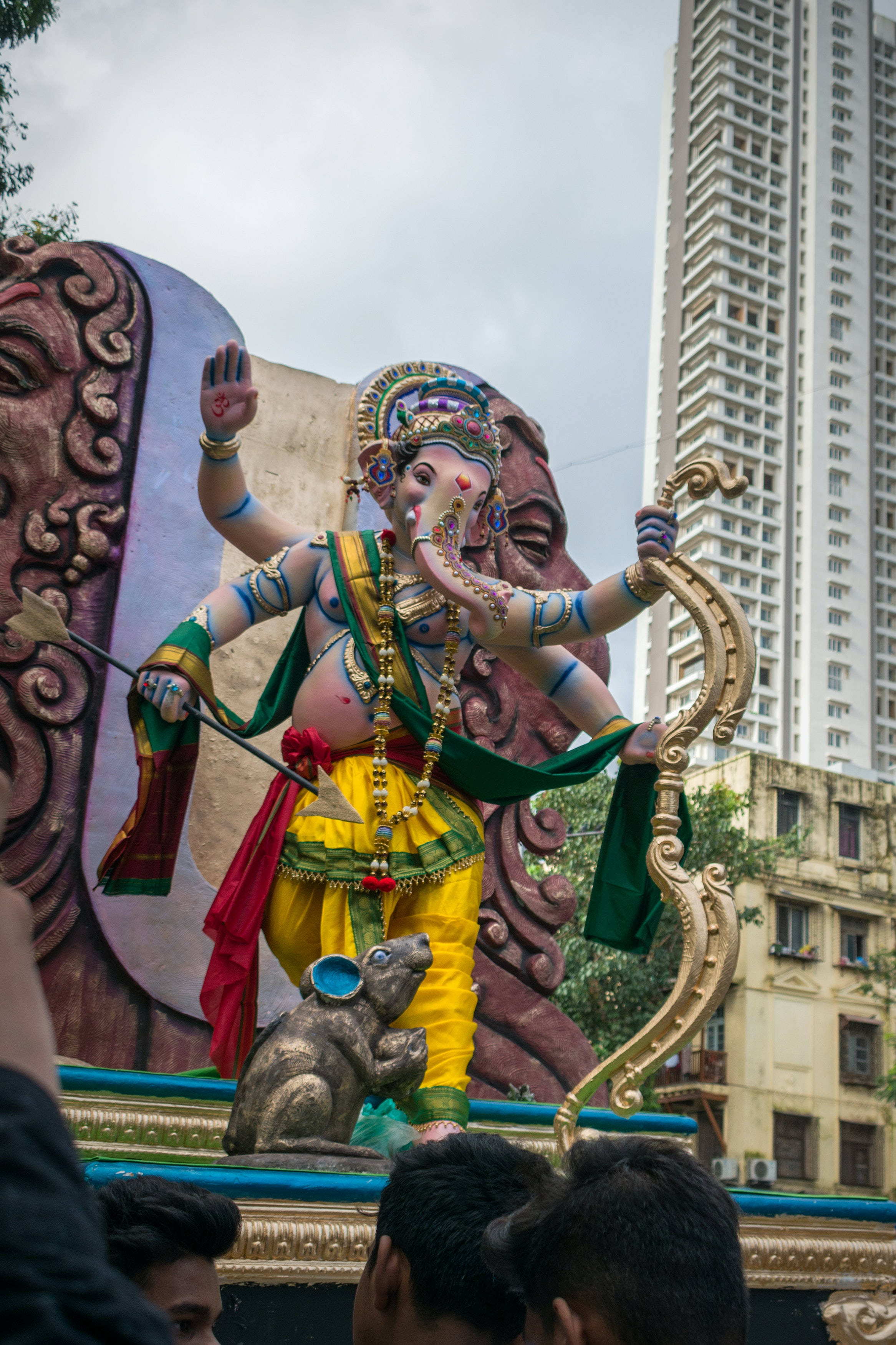 Ganesh 桌布 壁紙 — 一個色彩繽紛的象頭神像，擁有象頭和人類身體，站在節日期間裝飾的花車上，背景是現代城市的高樓大廈。