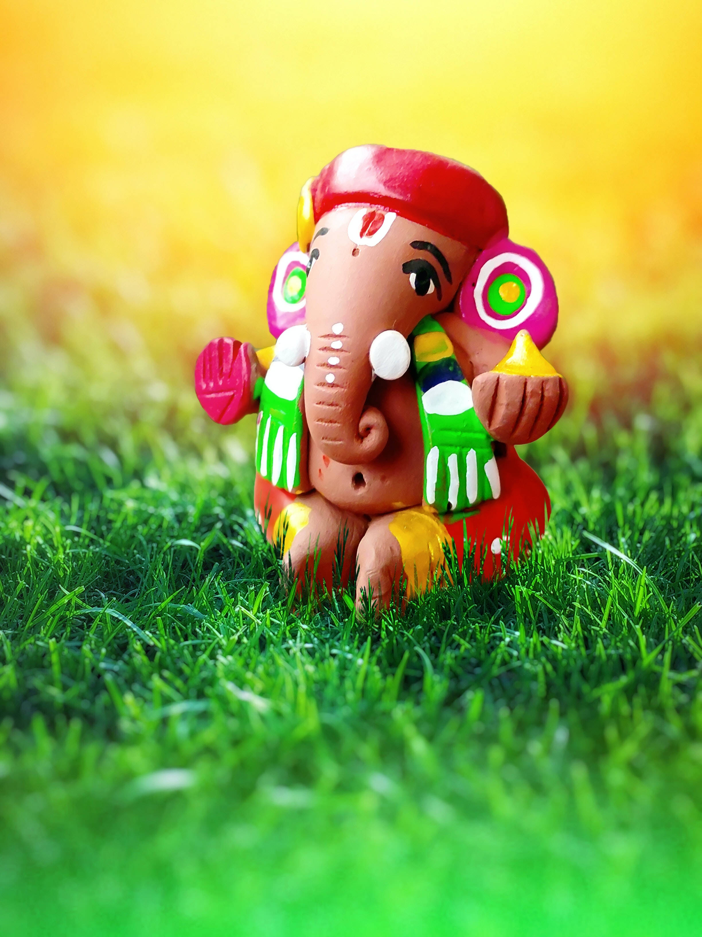 ganesh iPhone 桌布 壁紙 — 一個色彩鮮豔的彩繪粘土象頭神雕像，在溫暖的金色天空下，寧靜地坐在茂密的綠草地上。