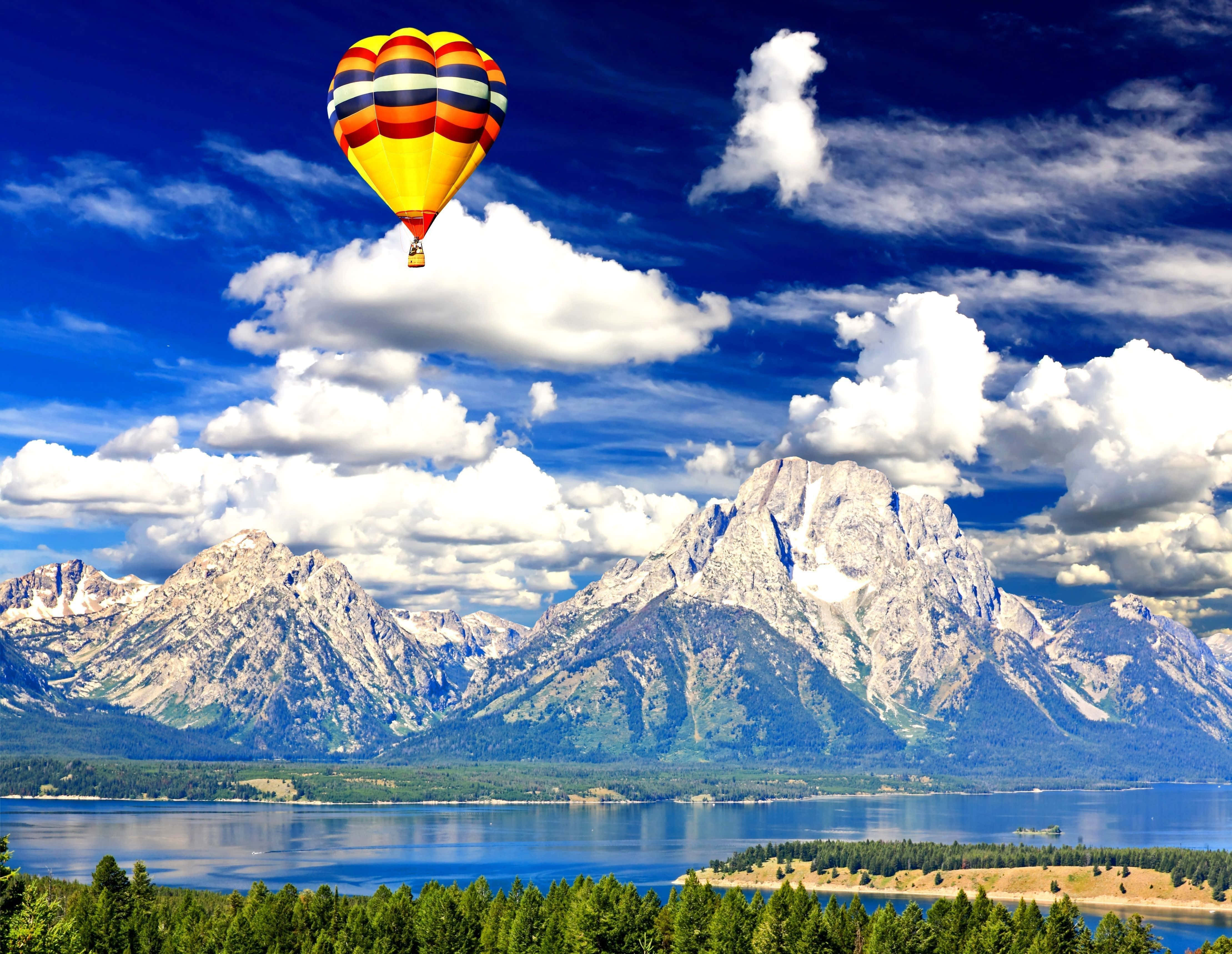 Hot Air Balloon 桌布 壁紙 — 一個色彩鮮豔的條紋熱氣球漂浮在寧靜的湖面上，背景是白雪覆蓋的山脈，天空湛藍，白雲朵朵。