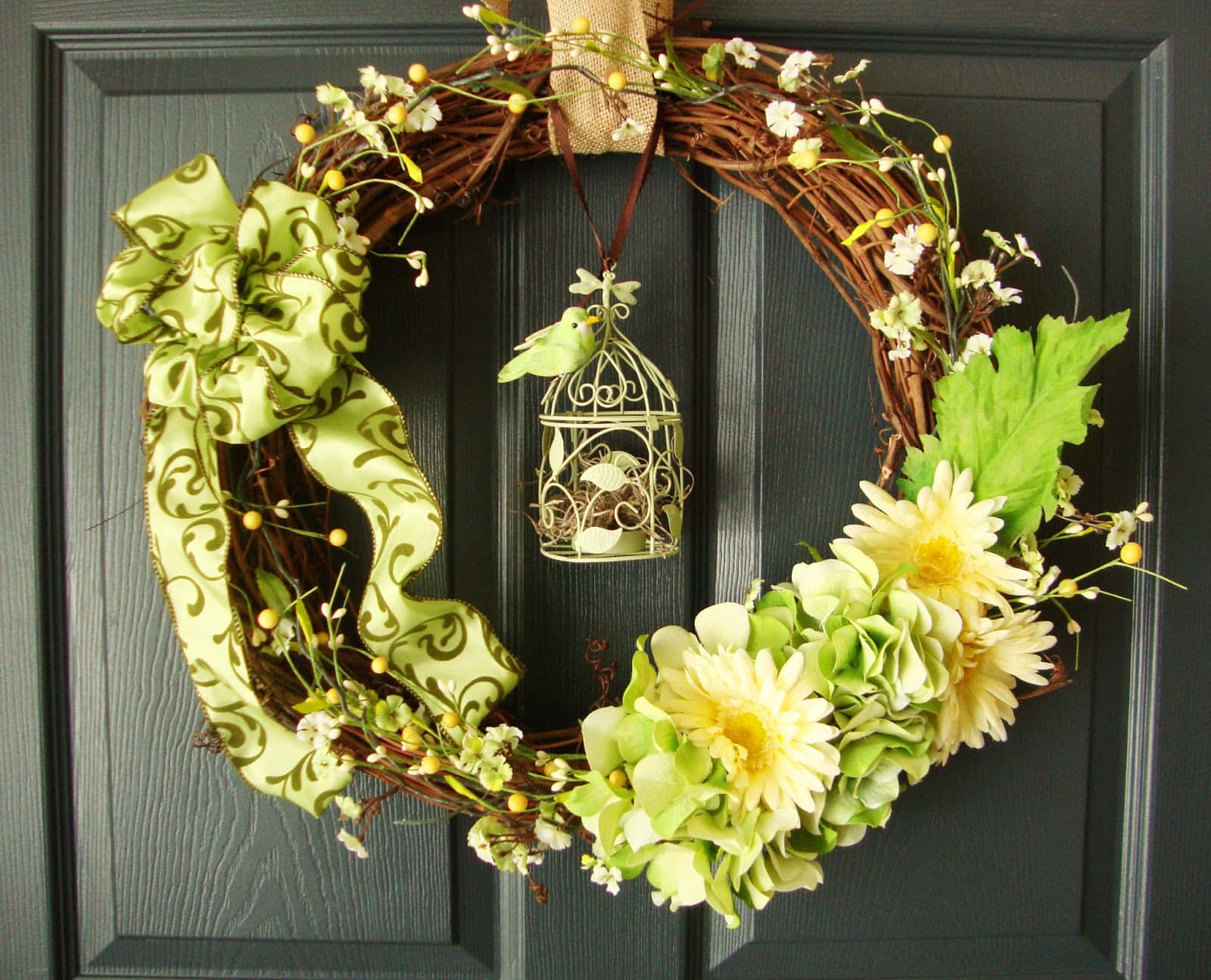 Spring Wreaths 桌布 壁紙 — 一個茂盛的綠色和黃色花卉花環，帶有裝飾蝴蝶結和鳥籠裝飾，掛在深灰色的木門上。