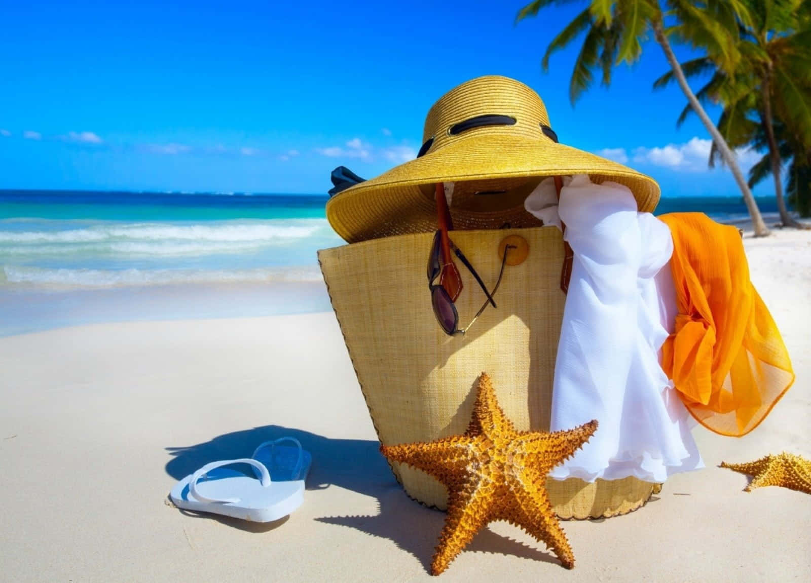 Beach Bag 桌布 壁紙 — 一個草編沙灘袋，內有太陽眼鏡和彩色圍巾，放在白色沙灘上，旁邊有海星和人字拖，棕櫚樹下。