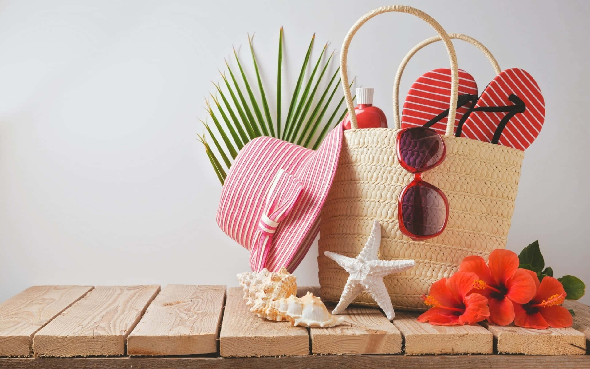 Beach Hat 桌布 壁紙 — 一個草編沙灘袋裝著紅色人字拖、太陽眼鏡和防曬霜，旁邊有粉紅色帽子、海星、貝殼和鮮豔的木芙蓉在木板上。