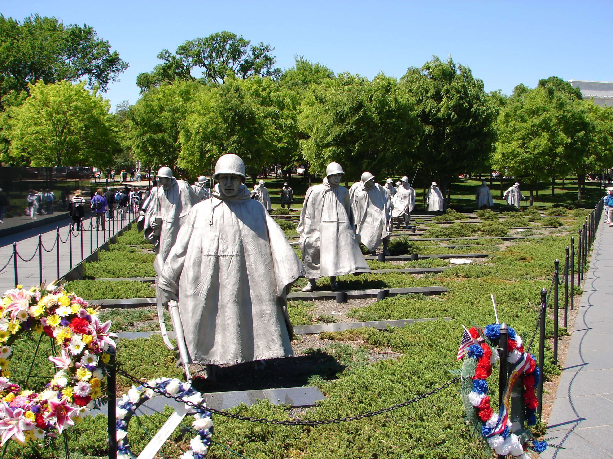 Memorial 桌布 壁紙 — 一個莊嚴的紀念碑，灰色士兵雕像在陽光明媚的藍天下穿過草地公園，底座上擺放著色彩繽紛的花圈。