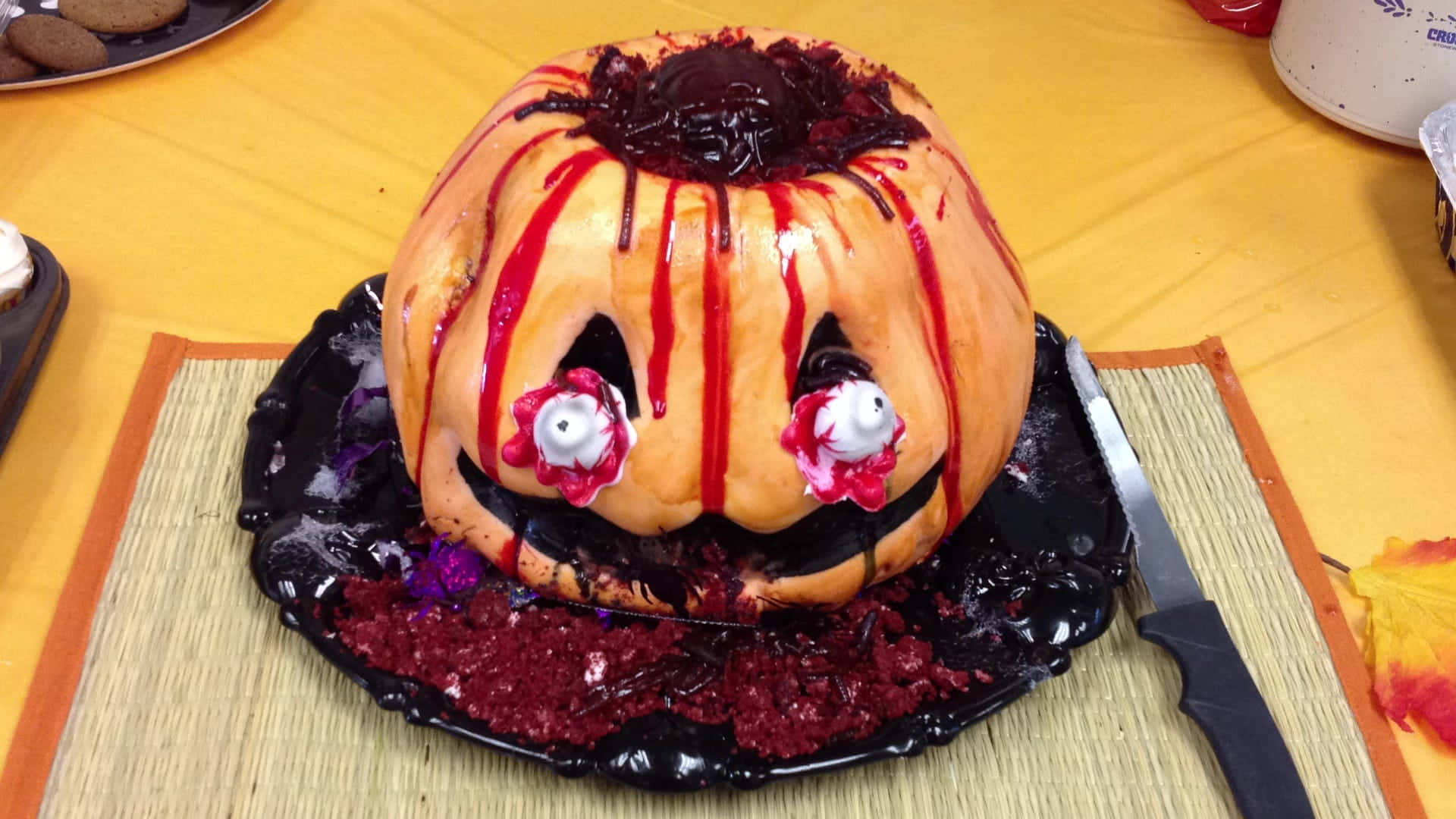 Halloween Cake 桌布 壁紙 — 一個萬聖節主題的南瓜形蛋糕，上面有紅色糖霜滴落和糖果眼睛，放在黑色盤子上。