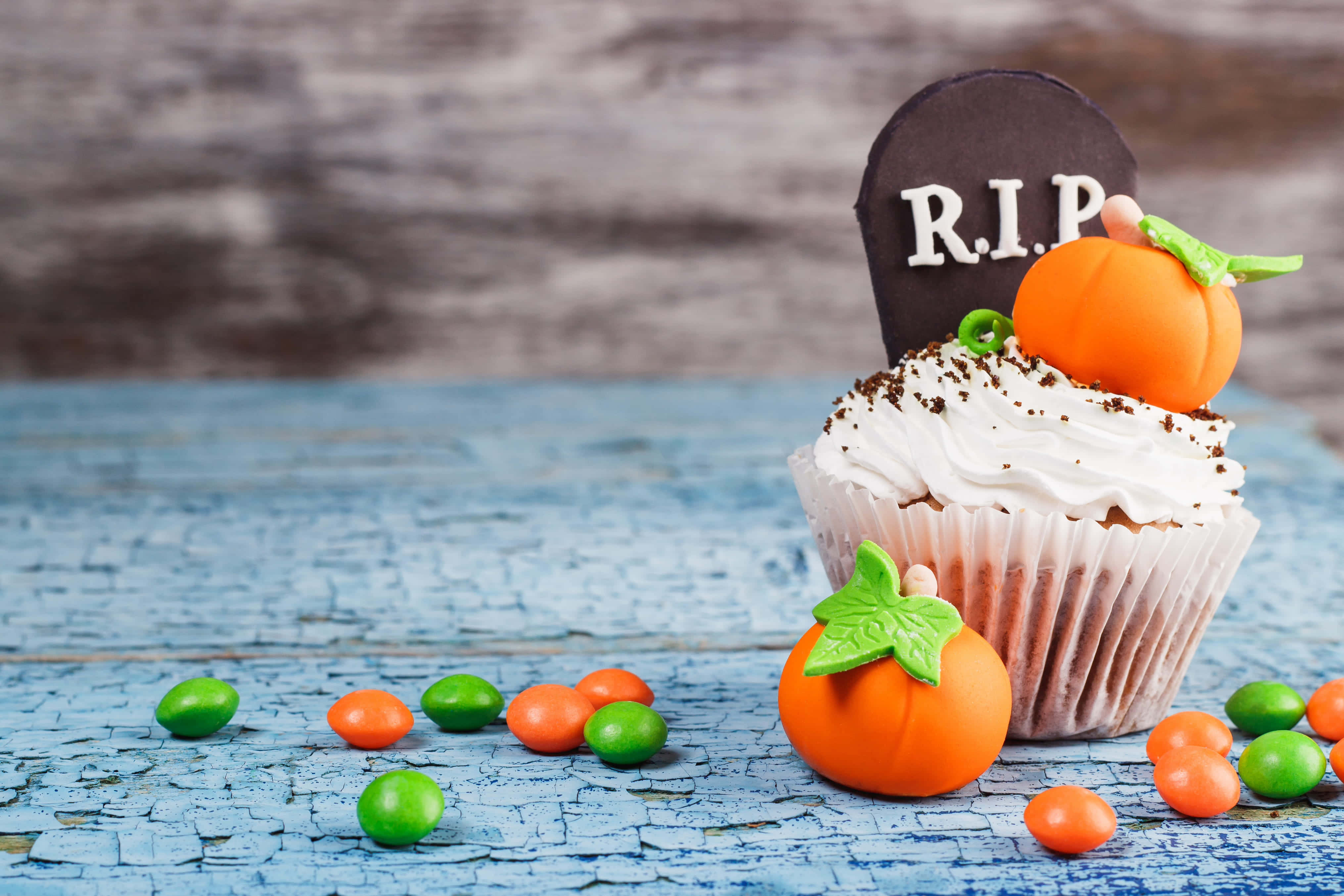 Halloween Cupcakes 桌布 壁紙 — 一個萬聖節紙杯蛋糕，上面有墓碑和南瓜裝飾，放在一個風化的藍色木質表面上，周圍環繞著糖果。