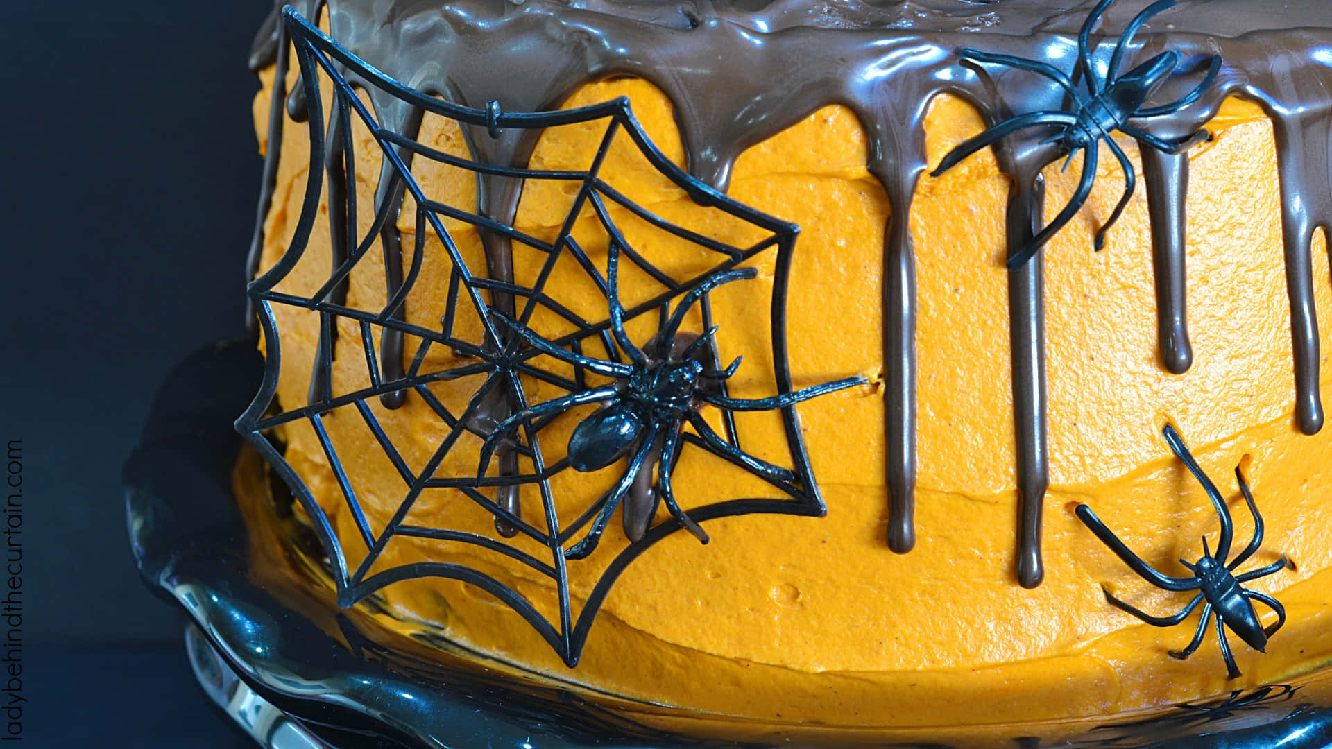 Halloween Cake 桌布 壁紙 — 一個萬聖節蛋糕，帶有橙色糖霜、朱古力滴和蜘蛛網上的黑色塑膠蜘蛛。