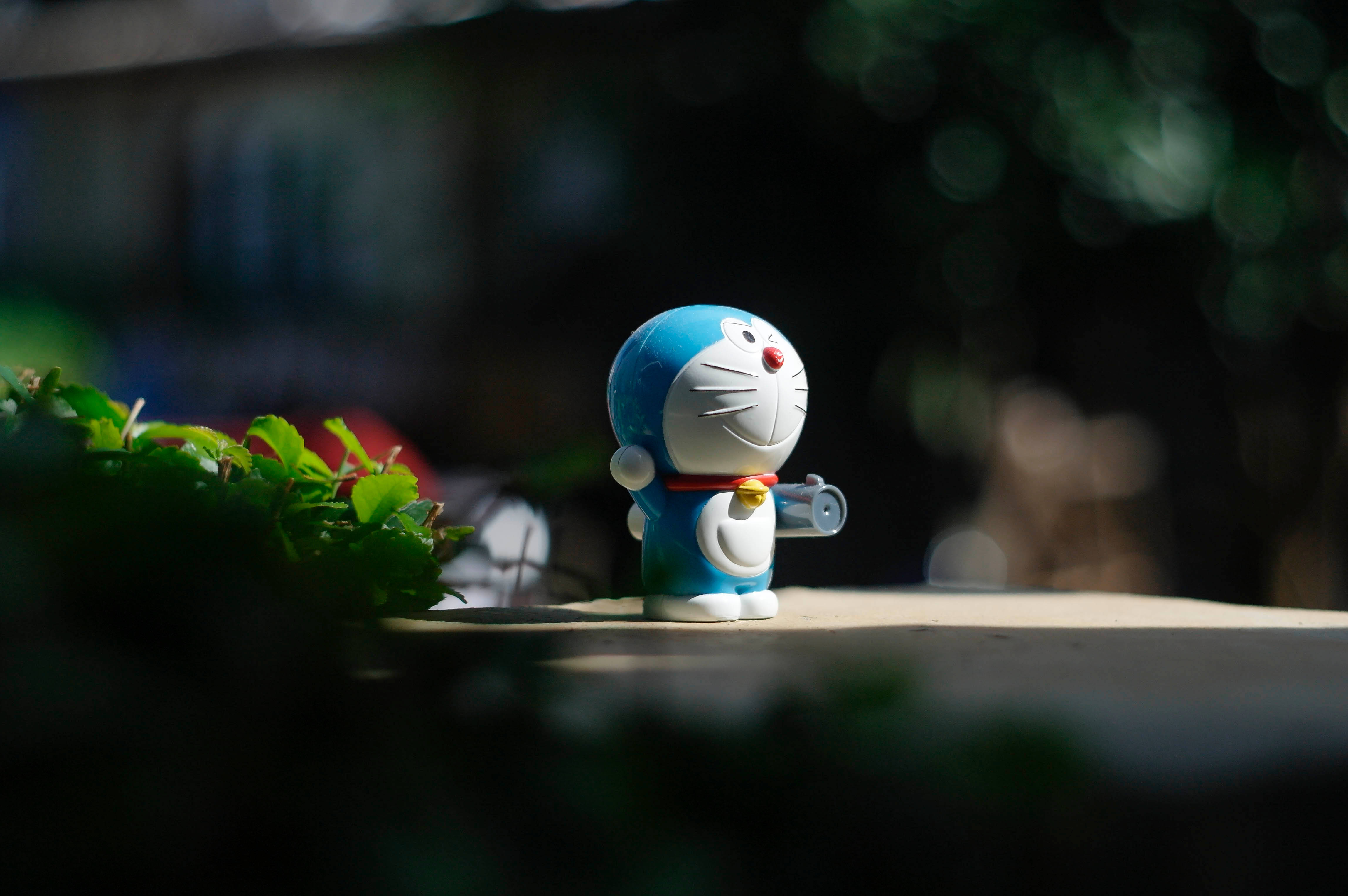 Doraemon 桌布 壁紙 — 一個藍白色嘅多啦A夢公仔企喺柔和陽光下，旁邊有綠色樹葉，背景係模糊嘅戶外景色。