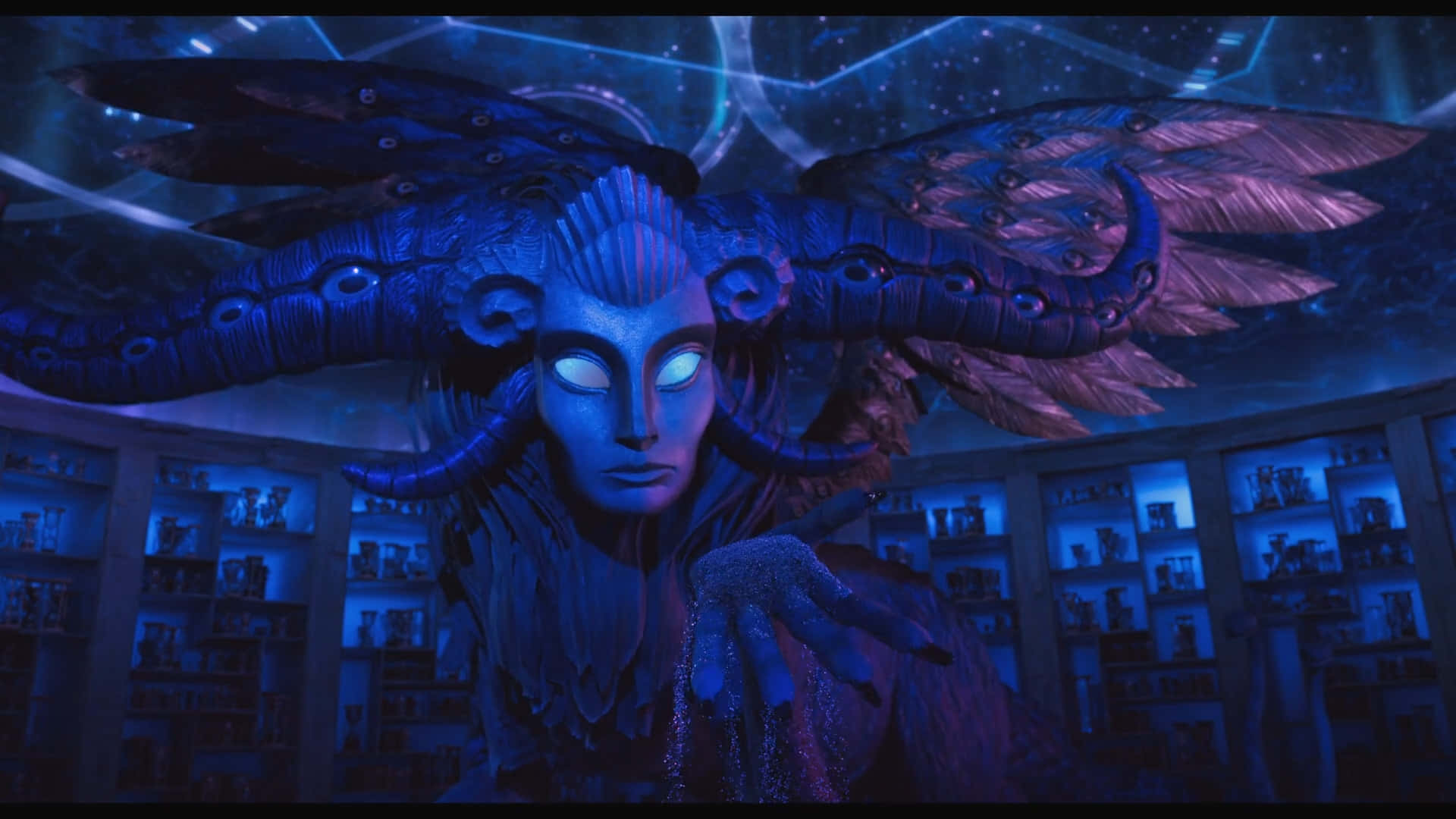 Guillermo Del Toro's Pinocchio 桌布 壁紙 — 一個藍皮膚、有翅膀、發光眼睛和華麗角的生物，站在一個昏暗的房間裡，房間裡擺滿了架子上的工藝品，天花板是星空。