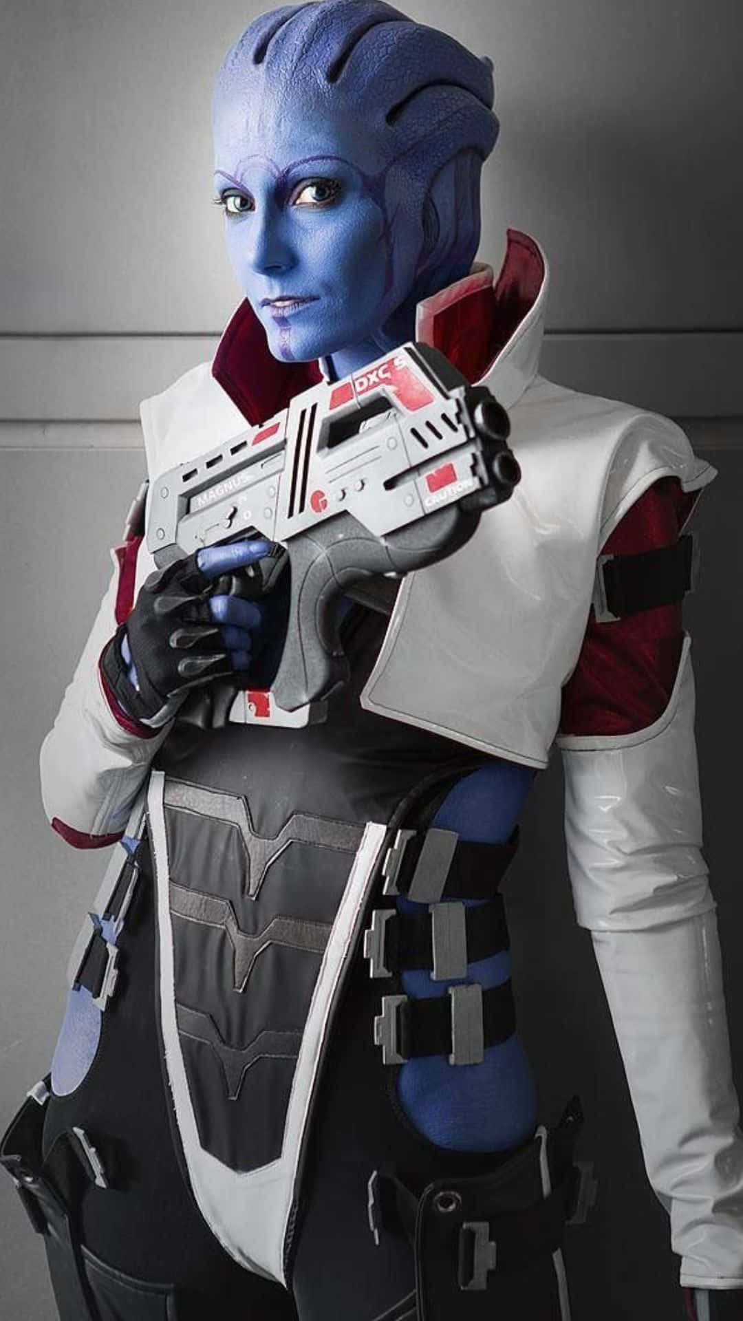 Aria T'loak 桌布 壁紙 — 一個藍皮膚外星人角色扮演者，身穿白色、紅色和黑色的未來風格服裝，手持一把細節豐富的科幻爆能槍，背景是純灰色牆壁。