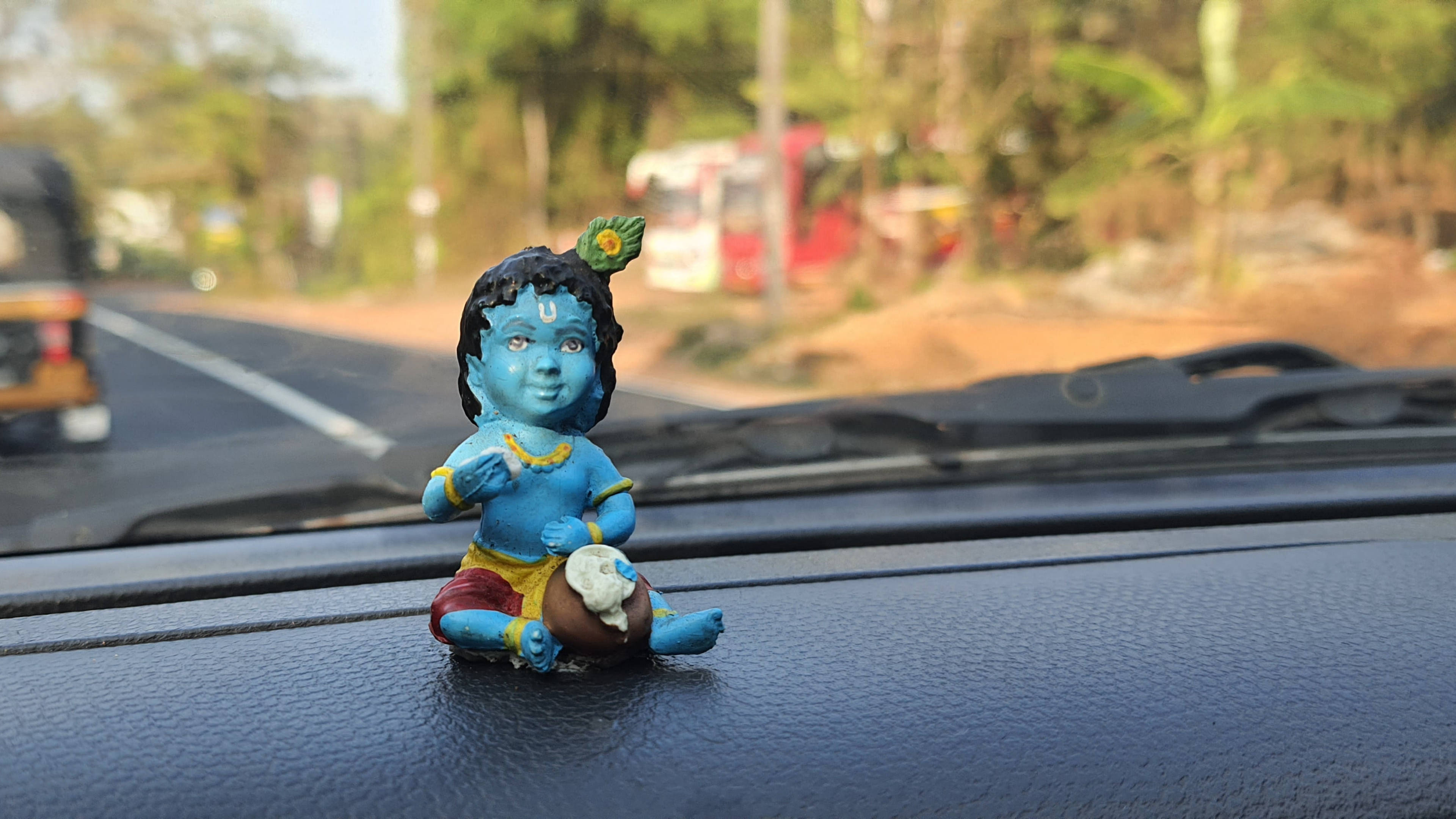 krishna ji 桌布 壁紙 — 一個藍皮膚的嬰兒克里希納雕像放在汽車儀表板上，背景是模糊的路邊景色。