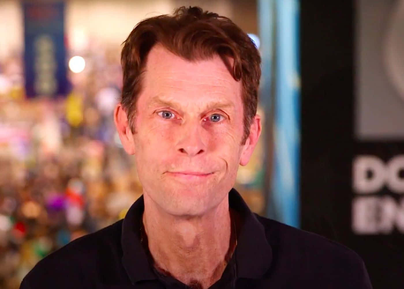 Kevin Conroy 桌布 壁紙 — 一個藍眼睛、棕色頭髮的男人，穿著黑色Polo衫，在模糊多彩的背景前微微微笑。