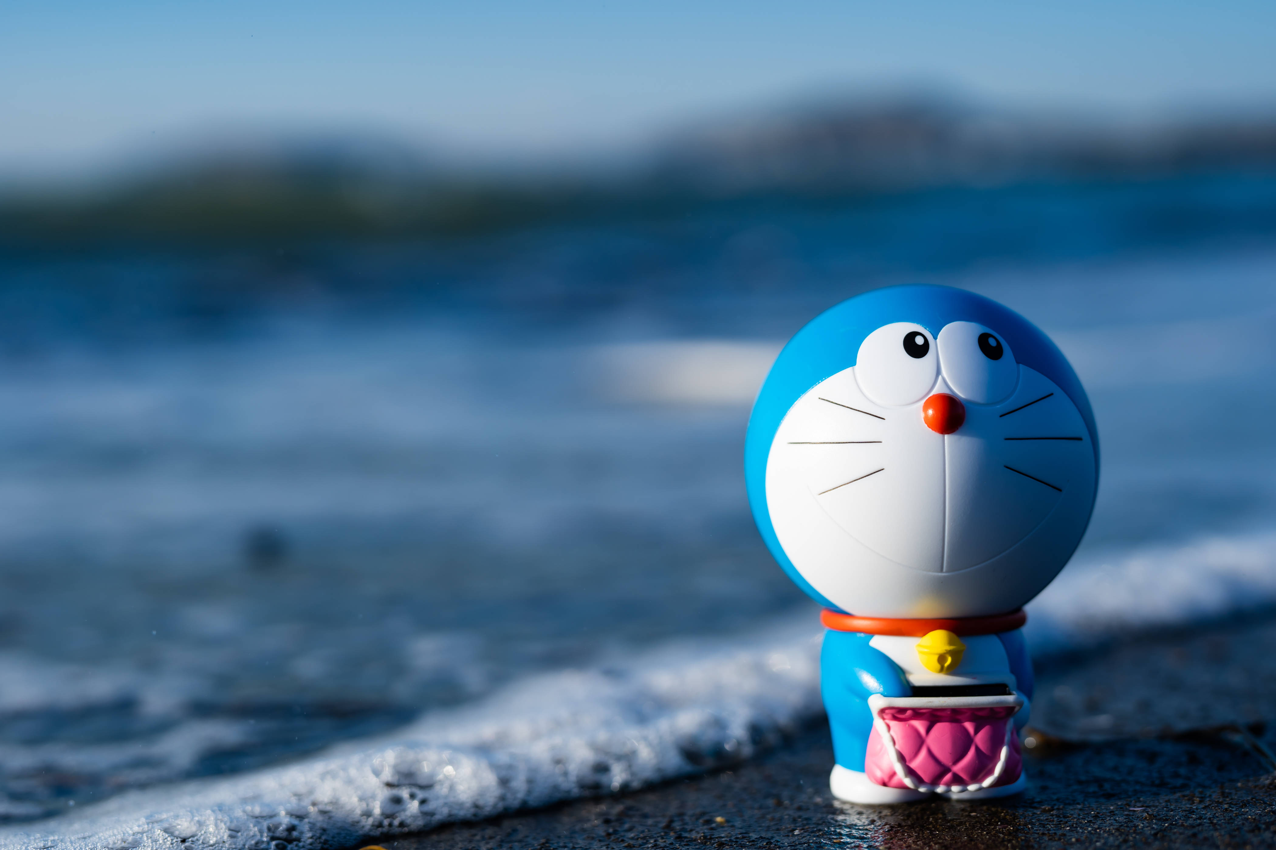 Doraemon 桌布 壁紙 — 一個藍色多啦A夢公仔企喺濕漉漉嘅沙灘上，溫柔嘅海浪喺晴朗藍天下緩緩湧入。
